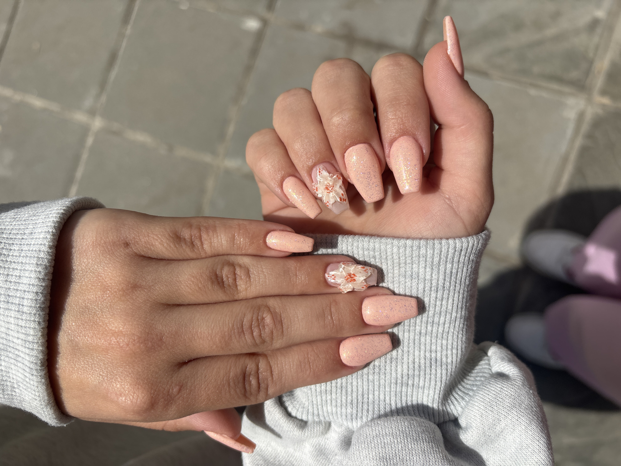The image shows a close-up of two hands with manicured nails. The nails are long, coffin-shaped, and painted a light pink color with glitter. The ring finger on each hand has a decorative floral design. The hands are resting on top of a light gray knitted sweater. The background appears to be a paved surface under sunlight. There are no visible landmarks to confirm the location as Sofia, Bulgaria.