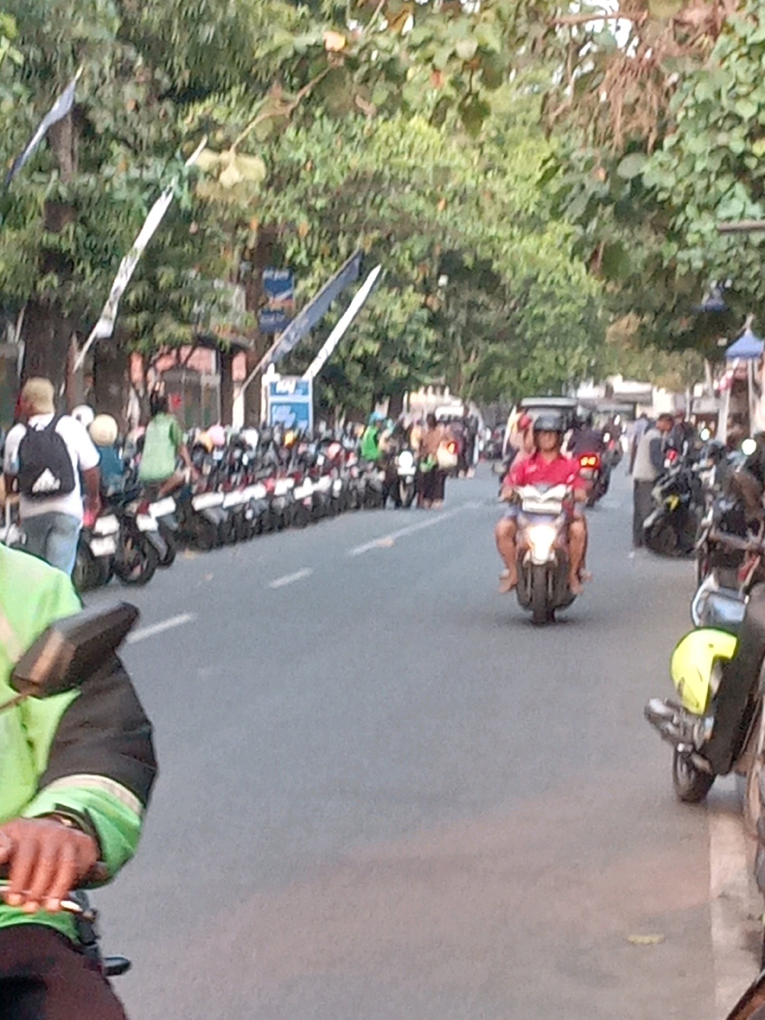 The image shows a street scene in Yogyakarta, Indonesia, with several motorcycles parked on the side and others in motion. In the foreground, a person wearing a green jacket is partially visible, holding onto a motorcycle's handlebars. Further down the street, a man in a red shirt and helmet is riding a motorcycle with a passenger. In the background, more motorcycles are lined up, and trees and buildings can be seen. The overall impression is of a busy urban environment with a prevalence of motorcycle transportation.