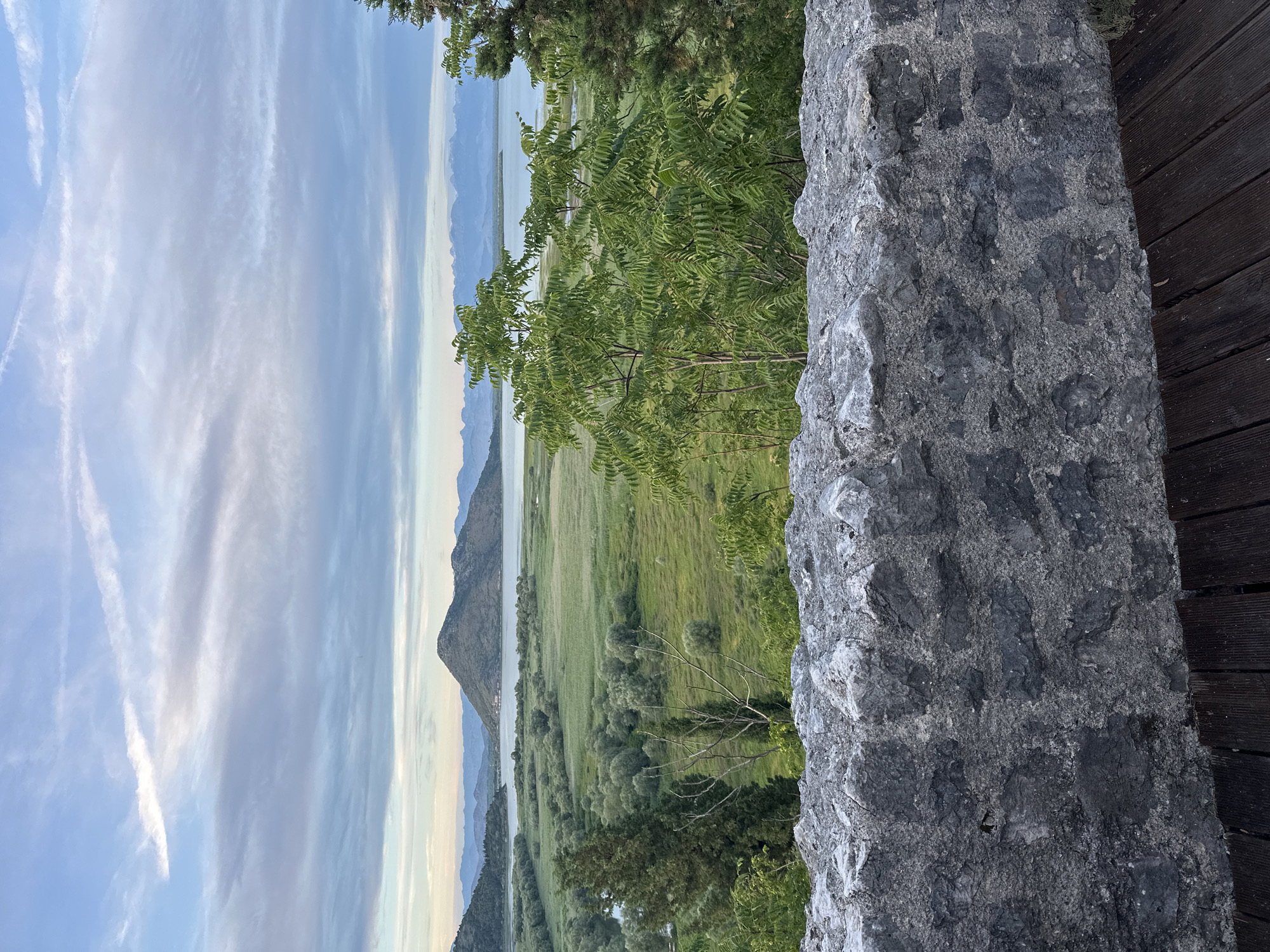 The image depicts a serene landscape viewed from an elevated position, likely a vantage point or balcony. In the foreground, a rustic, grey stone wall runs horizontally across the frame, with dark wooden planks visible beneath it. Beyond the wall, the scene opens up to a sprawling green valley, dotted with lush trees and vegetation. A calm body of water, possibly a lake or river, meanders through the landscape, reflecting the soft, overcast sky. In the distance, a prominent, conical mountain rises from the water, its silhouette softened by the atmospheric haze. A range of smaller, undulating hills forms a backdrop, fading into a pale blue sky streaked with wispy clouds. The overall impression is one of natural beauty and tranquility, suggesting a location in or around Virpazar, Montenegro, known for its picturesque views of Lake Skadar. There are no people or animals visible in the image.