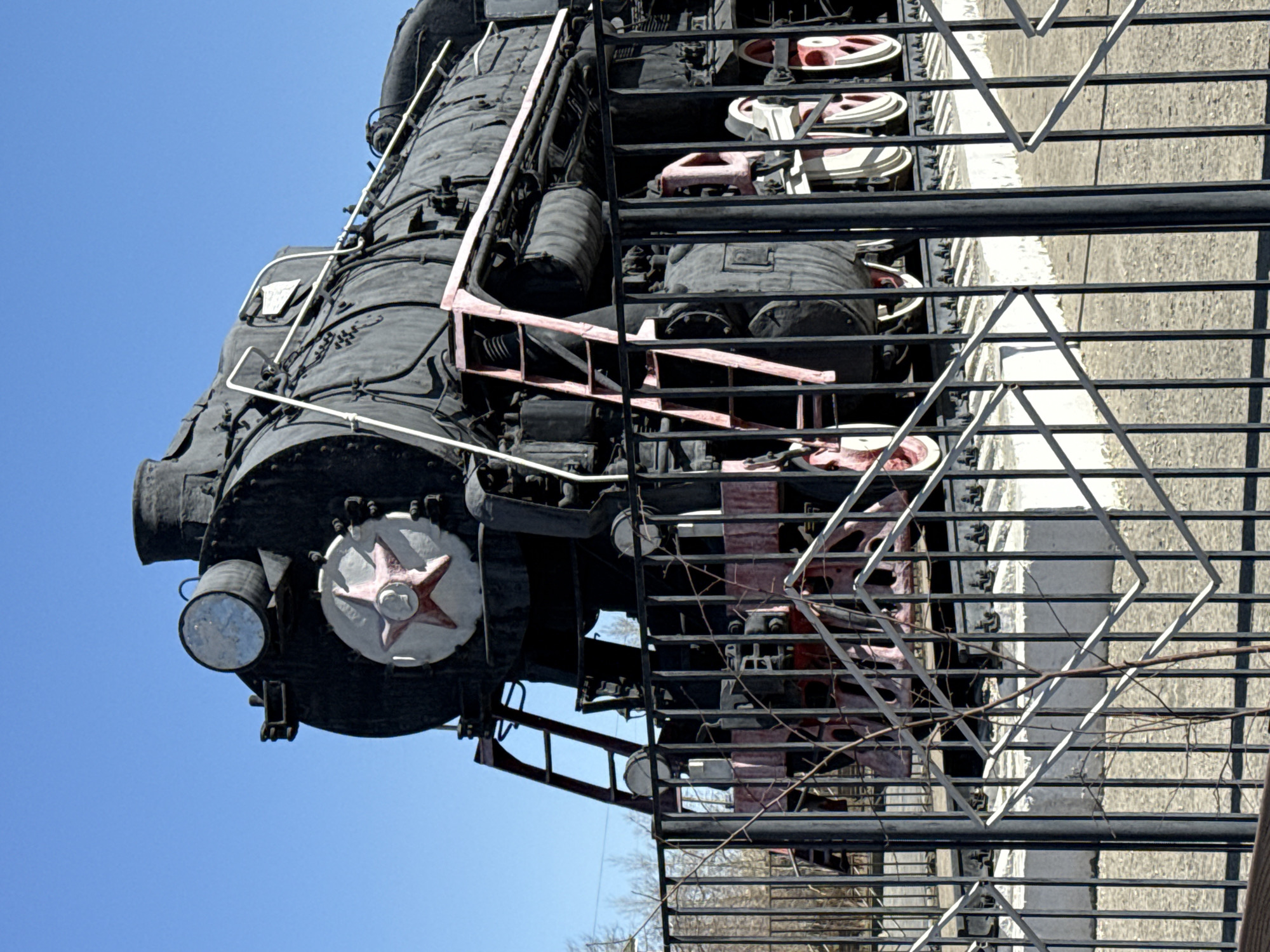 An antique locomotive is displayed outdoors in Borzya, Russia, viewed from a low angle against a clear blue sky. The locomotive features a black exterior with red and white accents, including a prominent red star on the front. It is partially obscured by a black metal fence with white diamond-shaped supports. The ground beneath the fence is visible, suggesting it is a paved surface. A few leafless tree branches are visible in the lower center of the frame.