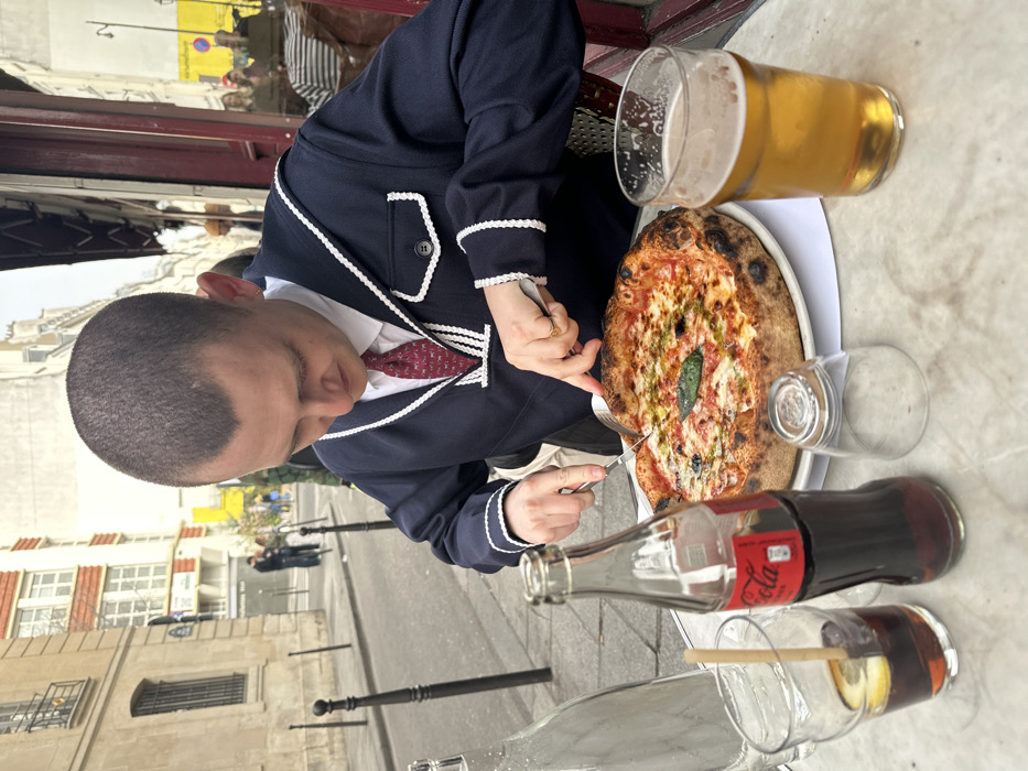 An adult male with a closely cropped buzz cut is seated at an outdoor cafe table, actively cutting a large Neapolitan-style pizza with a knife and fork. He wears a navy-blue sailor-style top with white trim, a white collared shirt, and a red patterned tie. A gold-colored ring is visible on his left ring finger.
The marble-patterned table holds the pizza on a white plate, a clear glass water bottle, a glass Coca-Cola bottle, a glass containing a dark beverage with ice, a lemon slice, and a wooden stirrer, an inverted empty glass, and a pint glass filled with a light amber liquid resembling beer.
The location is a street in Paris, France, characterized by multi-story Haussmannian-style buildings with light stone facades and dark windows. The building directly behind the male subject features a dark red-brown facade, indicative of a cafe or restaurant. Black bollards delineate the sidewalk. Blurred figures are visible on the street in the background. The scene is illuminated by natural daylight.