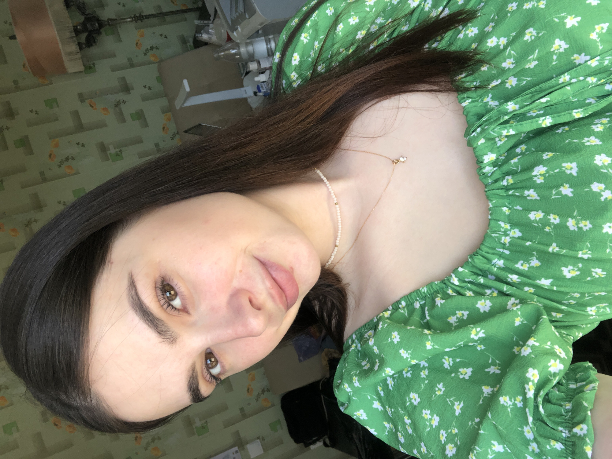 A young woman is depicted from a high-angle perspective. She has dark brown hair, light skin, and brown eyes. She is wearing a green dress with a small floral pattern. Around her neck, she wears a delicate necklace featuring small pearls and a pendant. The background consists of a wall with a geometric pattern and some equipment. It is not possible to determine if she is in the city of Noviy, Russia.