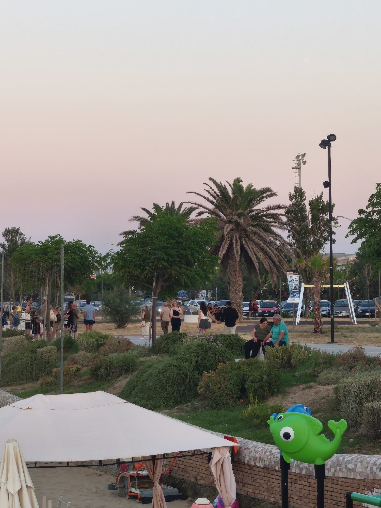 The image depicts a scene in the city of San Salvo, Italy, likely during the late afternoon or early evening, given the soft, pastel colors of the sky. In the foreground, a white canopy tent covers a sandy area, with a beige umbrella to its left. A child's playground element shaped like a green fish is visible on the right, mounted on a brick wall.
In the middle ground, a grassy slope is covered with various types of vegetation. Several people are gathered on this slope, some standing and others sitting. A couple, a man in a turquoise shirt and a woman in a dark green top, are seated together. Further back, a group of people are standing and conversing.
The background reveals a parking area filled with cars. Beyond the parking lot, there are trees, including palm trees, and buildings. Two tall light poles with multiple lamps are also prominent features in the scene, suggesting a public space like a park or a waterfront promenade. The overall atmosphere appears relaxed and social.