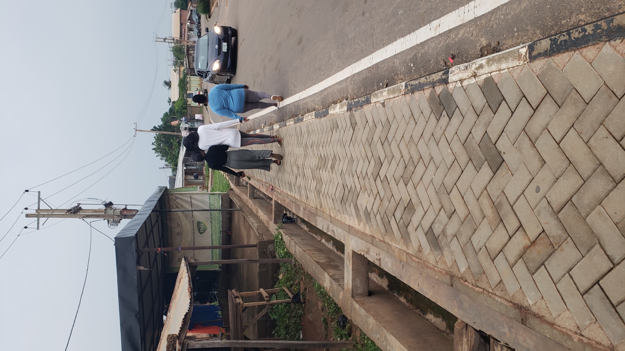 The image captures an everyday street scene in Narayi, Nigeria, with a slightly overcast sky. The primary subjects are three individuals walking away from the camera along a paved sidewalk, and a black car driving on the adjacent road.
In the foreground, a neatly paved sidewalk made of grey interlocked bricks in a herringbone pattern stretches from the lower right to the mid-left. Parallel to the sidewalk on its left runs a concrete drainage ditch. Further to the left of the ditch are various informal structures, possibly market stalls or temporary shelters, constructed with wooden frames, corrugated metal roofs, and green fabric tarpaulins. One of these tarpaulins has "TPM" visible in green lettering, along with partially visible words like "TRANSPARENT" and "EXCELLENCE" below it. Tall wooden utility poles with multiple power lines and a transformer are prominent on the far left, extending into the background.
Three people are walking along the pathway and road edge. The person closest to the road, on the right, wears a light blue top and dark bottoms, appearing to walk near or on the road's shoulder. Two other individuals are on the paved sidewalk: one in a white long-sleeved top and dark leggings, and another in a black top and grey pants. All three are facing away from the viewer.
To the right of the sidewalk is an asphalt road. A solid white line marks the edge of the road, and a broken black line runs along the curb. A black sedan, possibly a Toyota, is visible on the road, driving away from the camera. Its headlights are on, and a partial license plate "WJ204C" can be seen. In the background, more buildings and sparse vegetation are visible under the muted light of the overcast day.