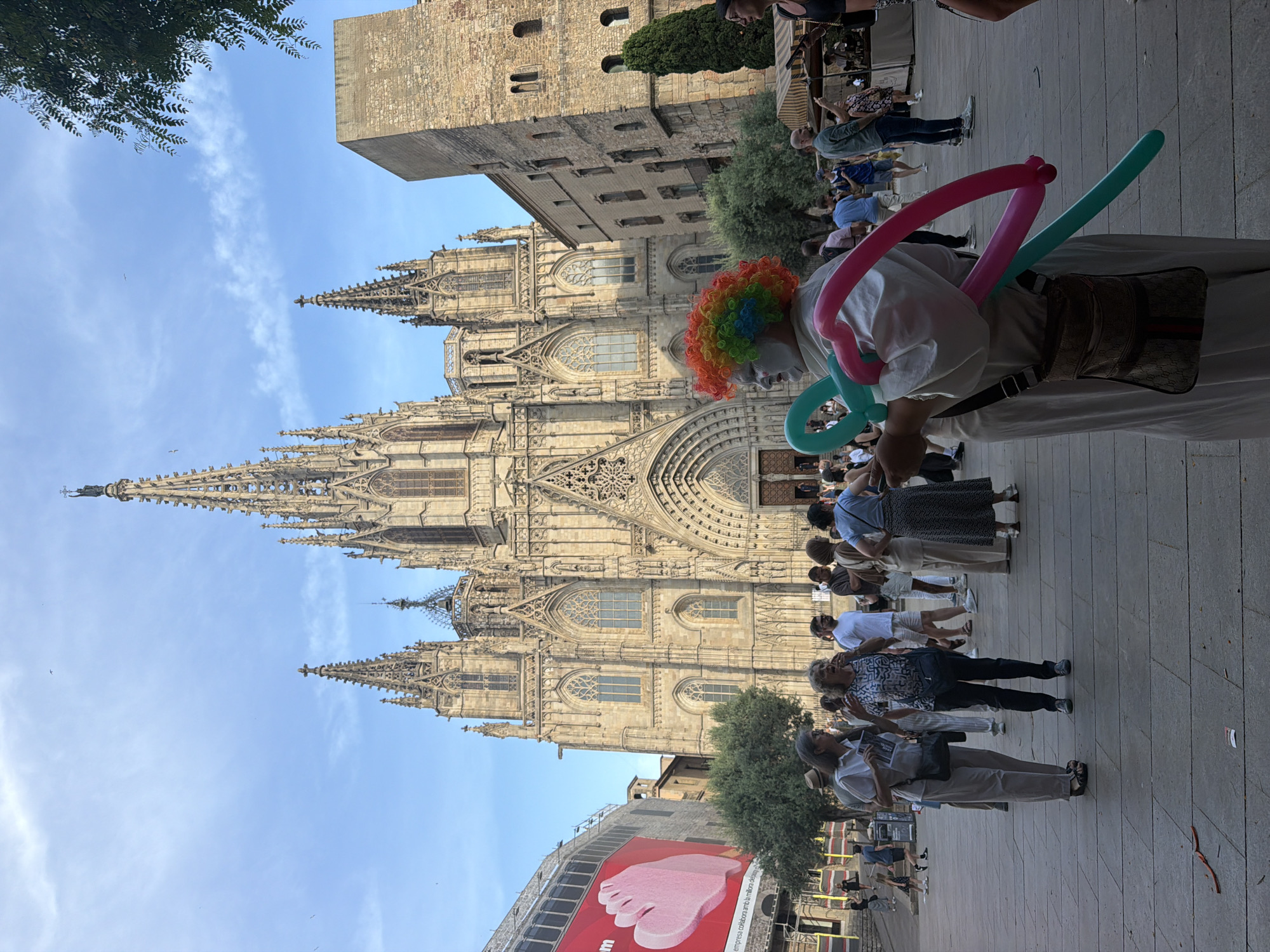 The image captures a scene in front of the Barcelona Cathedral, a Gothic Roman Catholic cathedral located in Barcelona, Spain. In the foreground, a person dressed as a clown, with brightly colored rainbow hair, white face paint, and holding pink and teal balloon animals, stands with their back mostly to the camera. They are wearing a white robe and a crossbody bag.
In the middle ground, a crowd of tourists and locals are gathered, some conversing and others observing the surroundings. Two women in the center are engaged in conversation. Behind them, a large, ornate Gothic cathedral with intricate spires and arched doorways dominates the background. The sky above is a clear blue with a few wispy clouds. To the right of the cathedral, a tall, stone building with arched windows is visible. The ground is paved with large, flat stones, typical of European city squares. A large billboard with a pink image of a foot is visible on the left side of the frame, indicating ongoing construction or advertising.