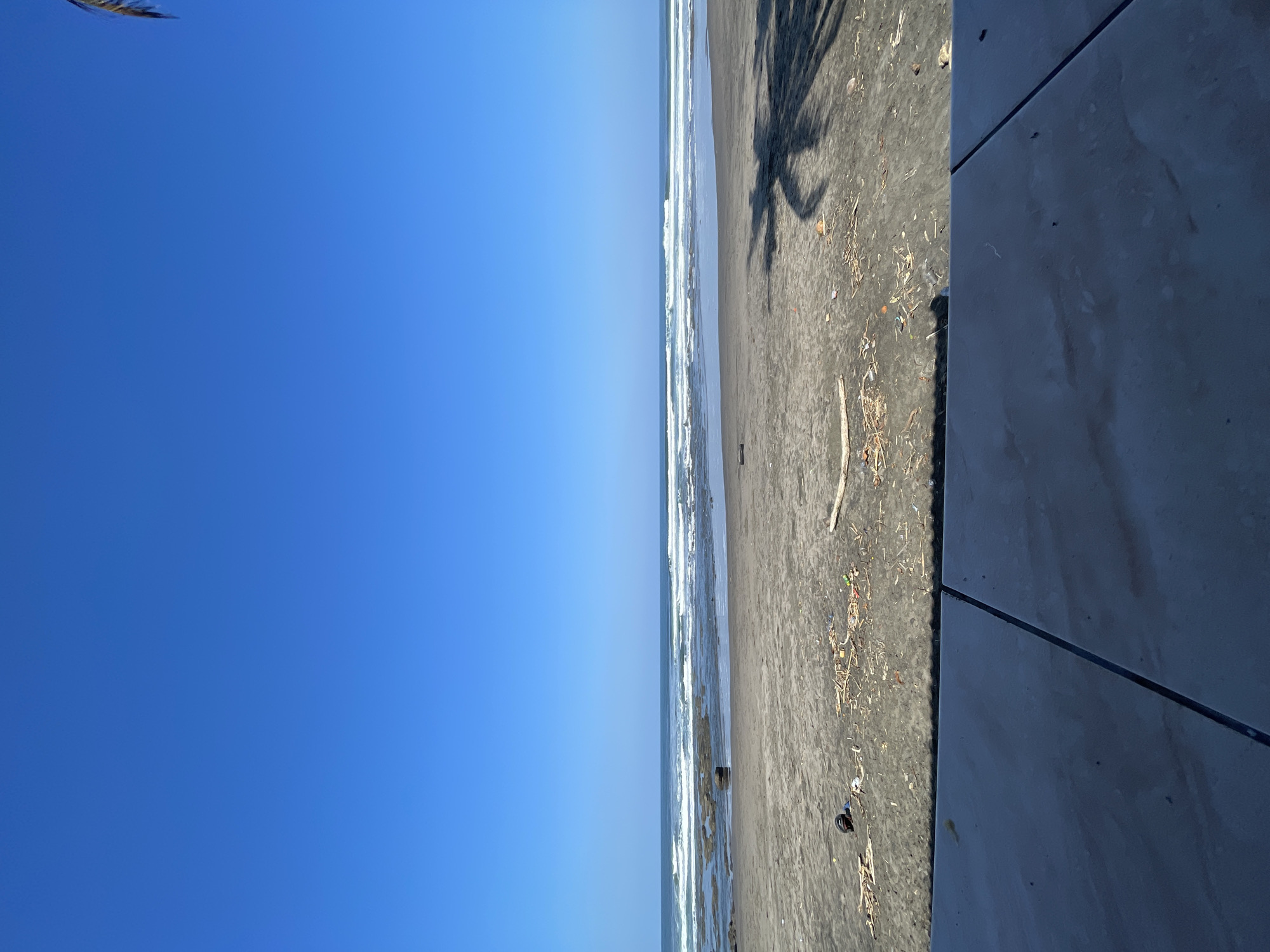 The image depicts a wide, expansive view of a beach on a clear, sunny day. The sky is a vibrant, cloudless blue, dominating the upper half of the frame. Below the sky, the ocean stretches across the horizon, with multiple lines of white waves breaking on a shallow reef or sandbar. The water appears calm between the breaking waves.
The beach itself is composed of dark, wet sand, with some areas appearing lighter and drier. Scattered across the sand are various debris, including driftwood, dried leaves, and what appear to be pieces of plastic or other refuse. A prominent shadow of a palm tree falls across the sand in the right portion of the image, suggesting the presence of palm trees nearby, likely just out of frame. In the mid-ground of the beach, there is a dark, rounded object that might be a rock or a piece of debris. To the left, closer to the water, a dark helmet lies on the sand.
In the foreground, a tiled surface with visible grout lines runs horizontally across the bottom of the image, suggesting the perspective is from a patio, walkway, or seating area overlooking the beach. The tiles are a muted, gray-blue color and appear to be slightly wet or polished.
The scene is tranquil and natural, characteristic of a tropical beach environment. The location is identified as the city of Pekutatan, Indonesia, which is known for its coastal areas and beaches. The overall impression is one of a serene, undeveloped beach scene under a bright sky.