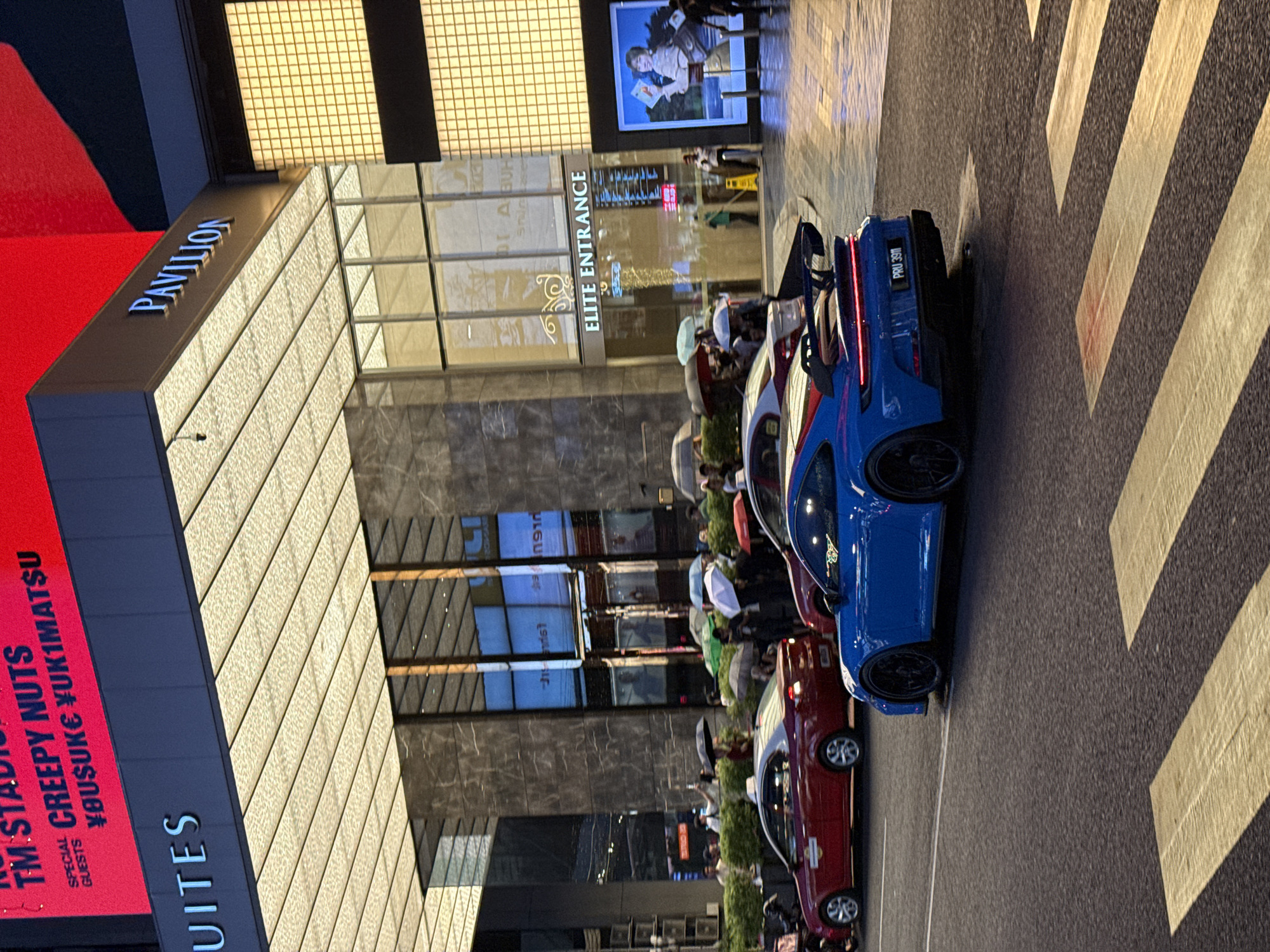 The image shows a street scene in Kuala Lumpur, Malaysia, in front of the Pavilion shopping mall. On the left, a red sign for TM Studios advertises "CREEPY NUTS" with special guests. The Pavilion's architecture is visible, including its entrance marked "ELITE ENTRANCE." A line of people with umbrellas extends from the entrance. Several cars, including a blue sports car with license plate "PRU 3911" and a red car, are parked on the street, which is marked with a pedestrian crosswalk.