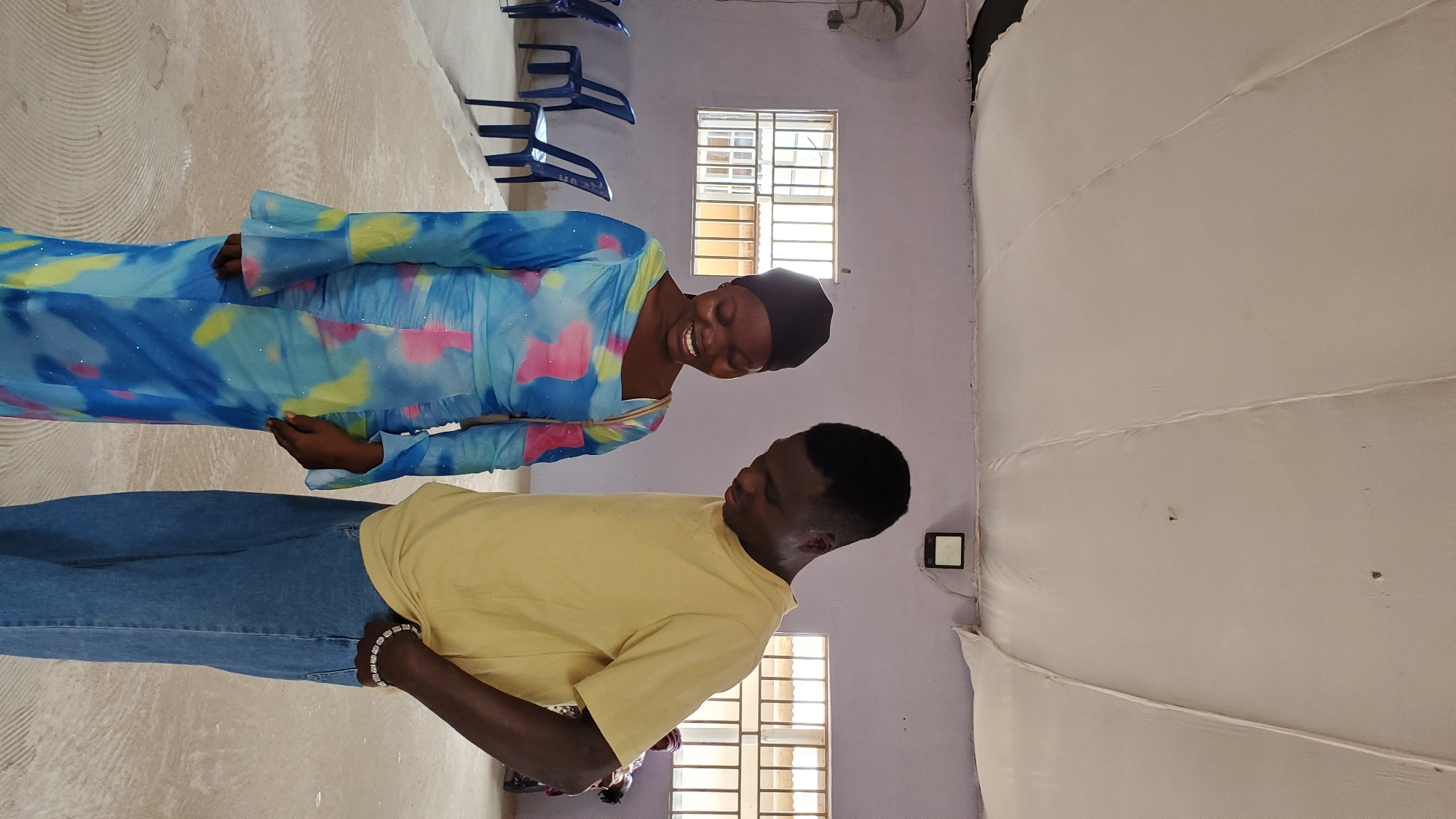 Two young adults, a woman and a man, are standing indoors, facing each other and looking down. The woman, on the left, is wearing a black headscarf and a vibrant blue, yellow, and pink tie-dye dress with flared sleeves. She is smiling broadly. The man, on the right, is wearing a light yellow t-shirt and blue jeans. He has a bracelet on his left wrist, which is tucked into his jean pocket. They appear to be in a large room with a white, fabric-draped ceiling. On the wall behind them are two windows with horizontal bars and a light-colored structure on the wall. In the background, blue chairs are arranged in rows. The floor is a light brown color with a circular pattern. The setting appears to be a community hall or a meeting space within Jalingo, Nigeria.