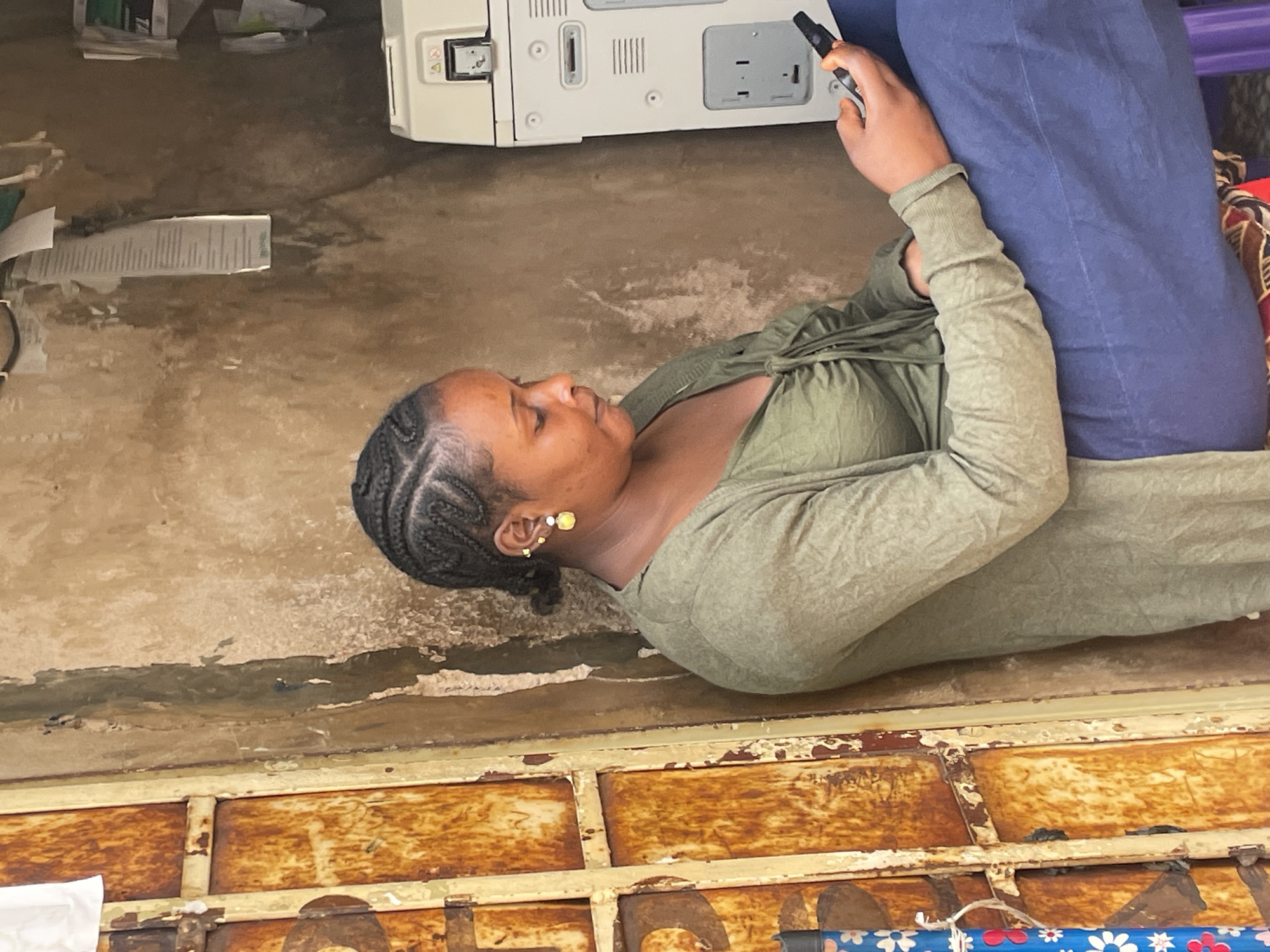 A woman lies on her side on a concrete floor next to a printer, holding a black object in her hand, presumably a cell phone. She wears an olive green shirt and blue jeans, and her hair is braided. To her left is a metal grate with peeling paint. The location is Jos, Nigeria.