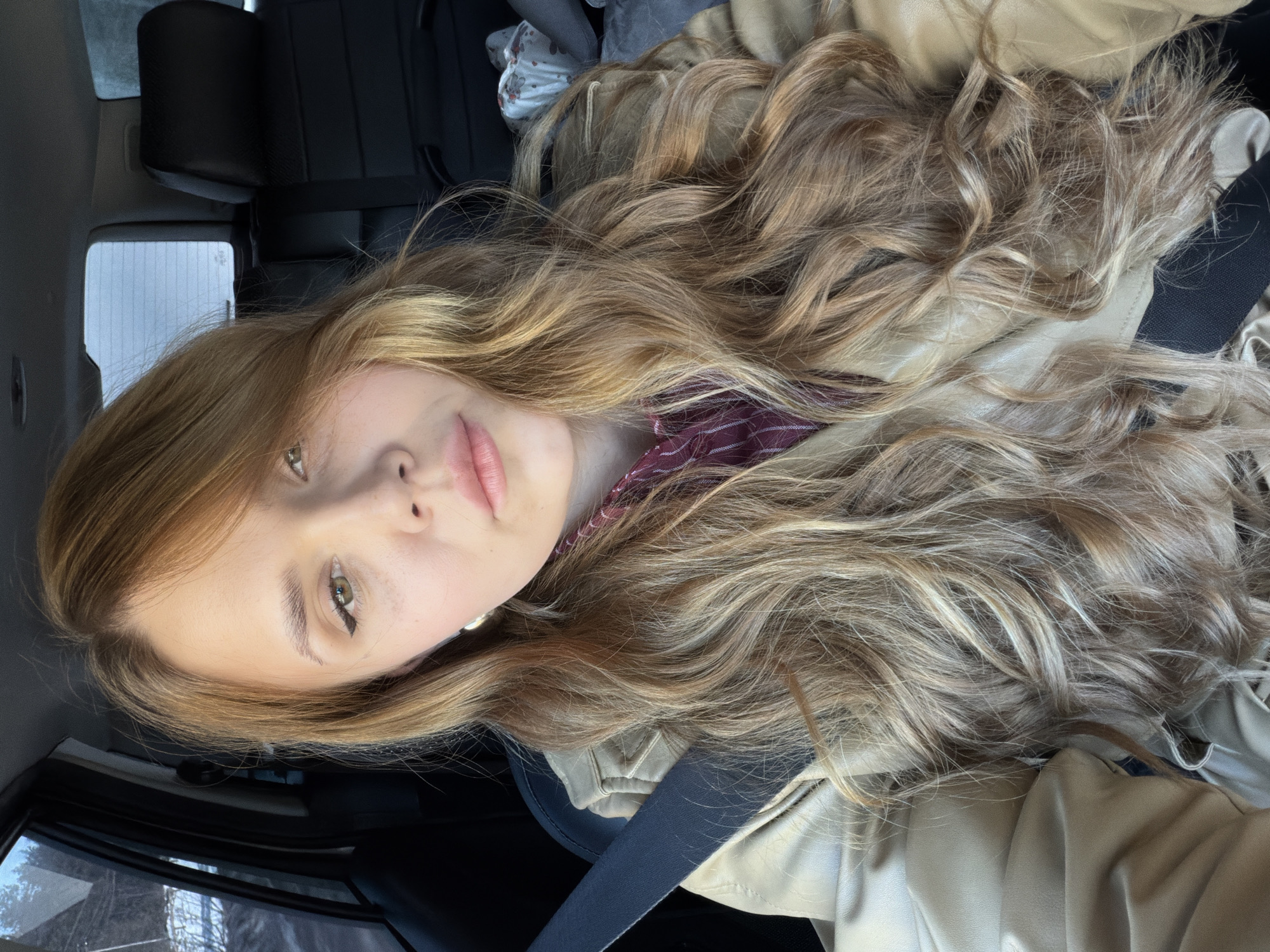 A young woman with long, wavy blonde hair sits in a car, wearing a tan jacket and a maroon and white striped shirt. She is wearing a seatbelt. The background shows the car interior, including the black seats and a glimpse of the outside through the car window. Location: Unknown, does not include Noviy, Russia.