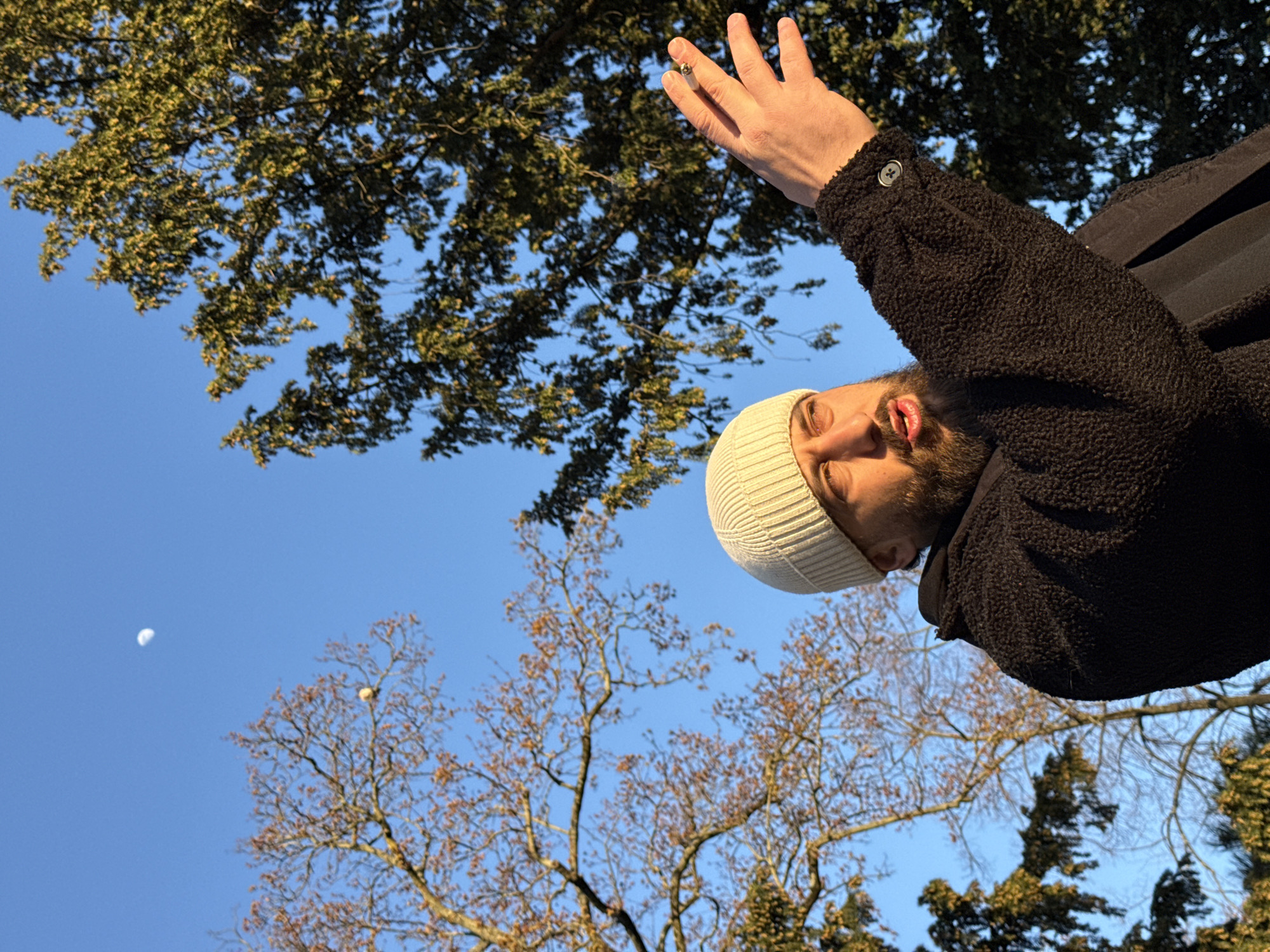 An outdoor scene in Strasbourg, France, depicts an adult male from the chest up, framed against a clear blue sky with a crescent moon visible in the upper left quadrant. The man, positioned on the right side of the frame, wears a cream-colored knitted beanie and a dark, textured fleece jacket. He has a beard, his mouth is slightly open, and his gaze is directed upwards and to the left. His right arm is extended upwards and to the right, with an open hand holding a lit cigarette between his index and middle fingers. The sleeve of his jacket features a dark button on the cuff. Two trees are prominent: a deciduous tree with sparse, reddish-brown leaves on the left, and a dense evergreen tree with dark green foliage on the right. A small, light-colored spherical object is lodged within the branches of the deciduous tree on the left. The scene is illuminated by warm, directional sunlight, indicating either sunrise or sunset.