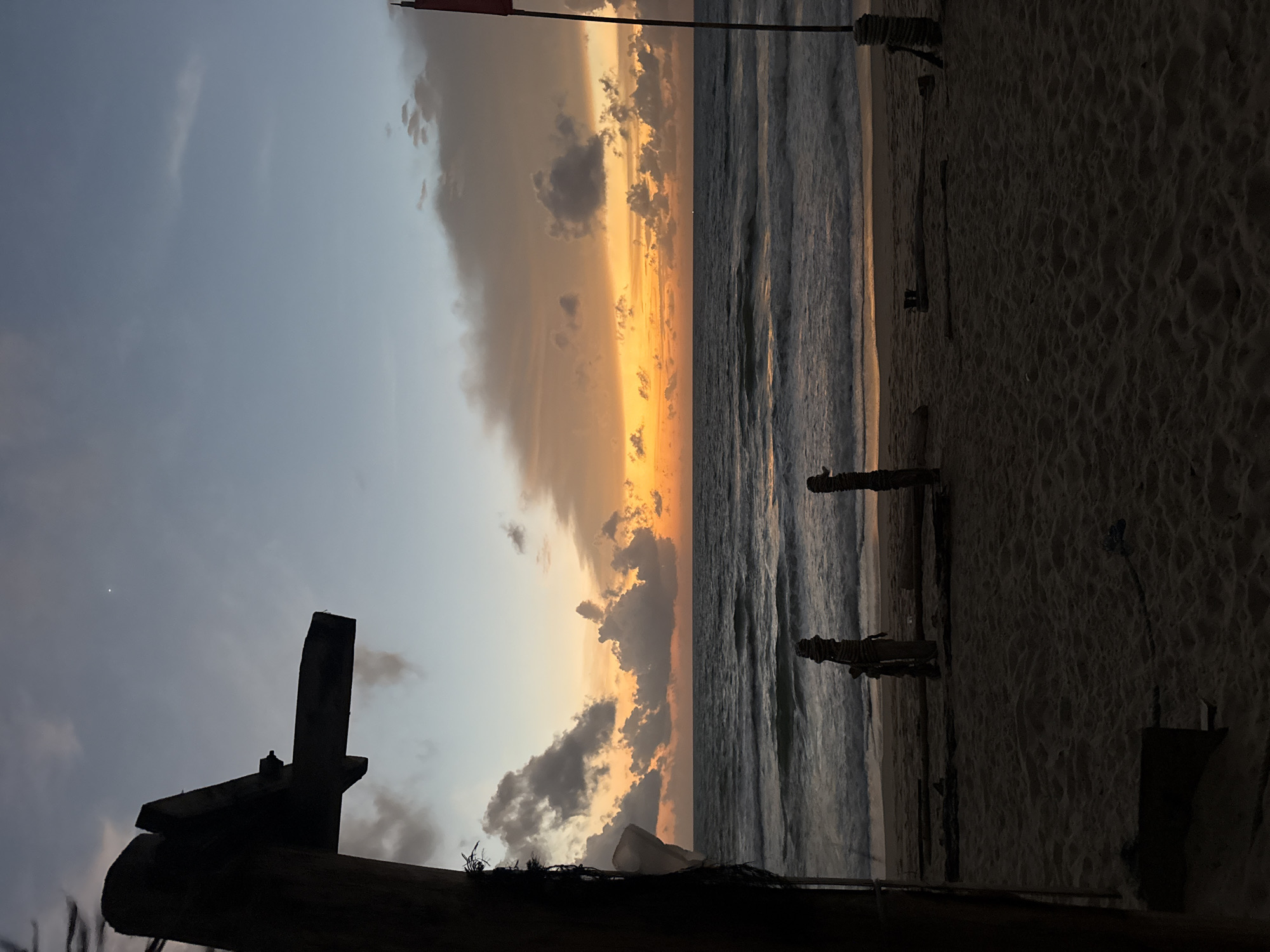 A serene beach scene unfolds at dusk in Choeng Thale, Thailand. The sky, transitioning from a pale blue to a vibrant orange and yellow gradient, showcases scattered clouds tinged with the setting sun's glow. In the foreground, a silhouetted wooden structure, possibly part of a shelter or pier, juts into the frame from the left. A red flag on a pole stands to the right, its dark silhouette against the colorful sky. The sandy beach is sparsely decorated with pieces of driftwood and two wooden posts wrapped with rope. Gentle waves lap the shore of the ocean, their crests catching the fading light. The overall atmosphere is tranquil, capturing the quiet beauty of a tropical sunset.