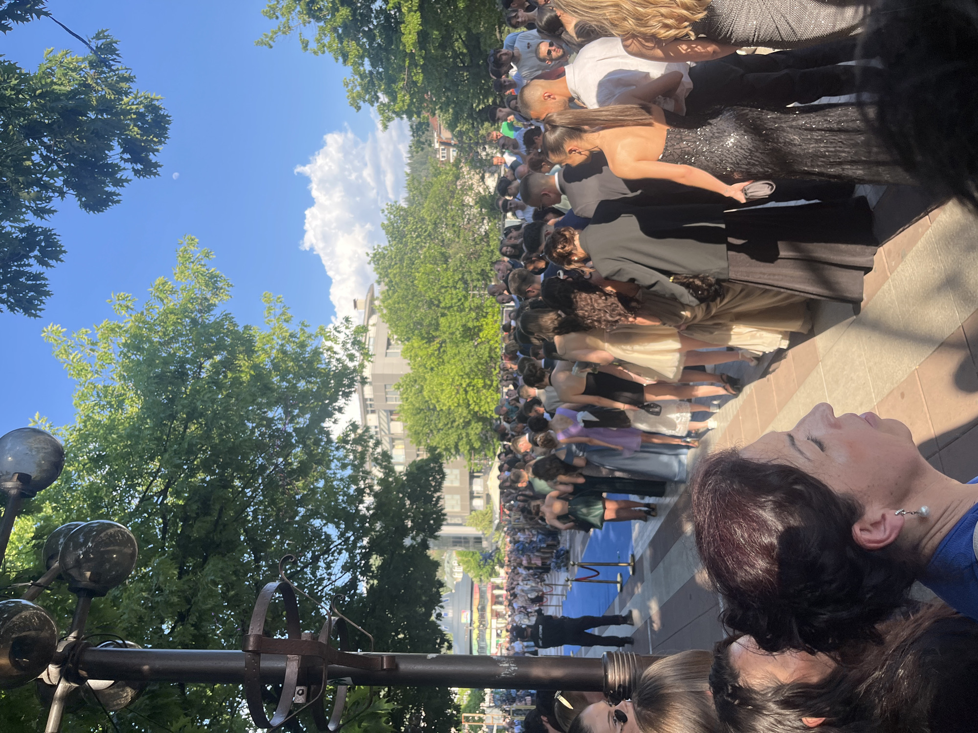 The image shows a crowd of people lined up on a sidewalk, possibly for an event or premiere, as suggested by the blue carpet and stanchions. Many individuals are dressed in formal attire, including evening gowns and suits. A portion of the crowd is turned toward the camera, with individuals in the foreground and background. In the background, there is a multi-story building. The photo has a high angle, showing trees, blue sky, and white clouds. The location is likely Blagoevgrad, Bulgaria, because of the architectural style and location.
