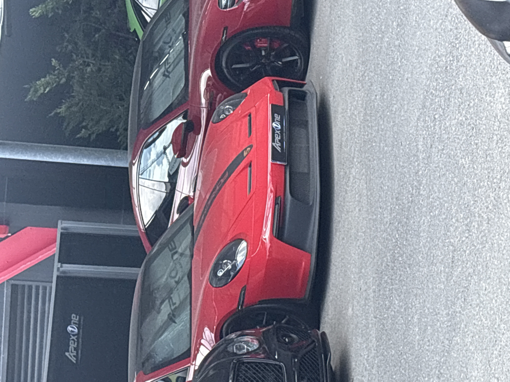 The image captures a street scene in Sofia, Bulgaria. Two red Porsche cars, each bearing the "Apex One" logo, are parked next to a building with "Apex One" signage. The ground is asphalt. In the background, there is greenery and a support pole. A black Bentley grille is partially visible under one of the Porsches.