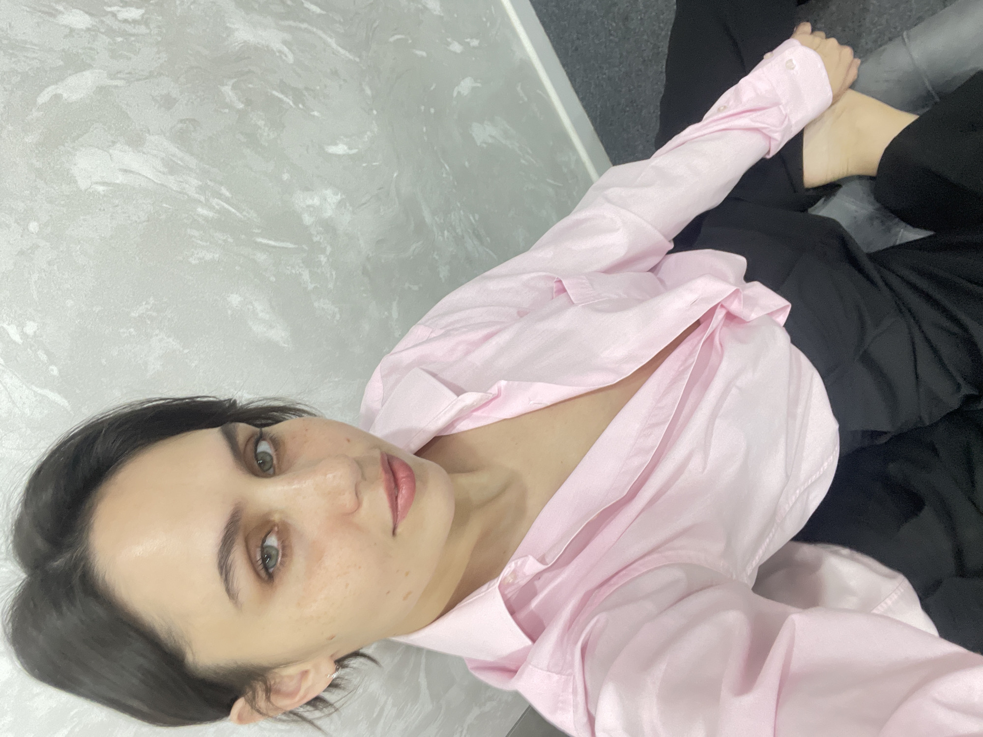 A woman with dark hair is taking a selfie. She is wearing a light pink button-down shirt and black pants. The background consists of a textured wall, likely indoors, and a dark surface, possibly a carpet. There is no specific information to definitively place this scene in Izhevsk, Russia.