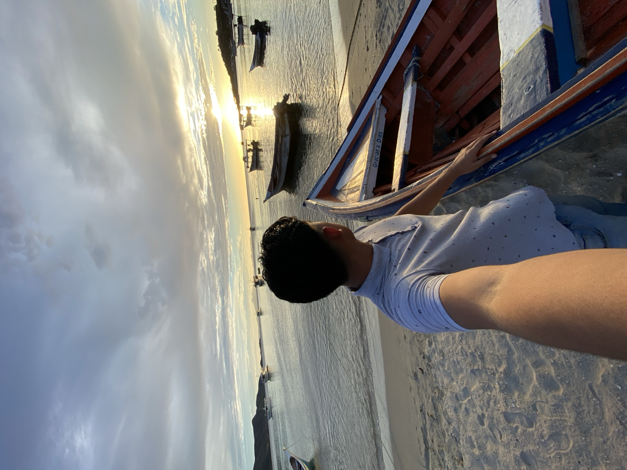 The image depicts a man standing on a sandy beach, looking out at a bay filled with boats. The sun is setting, casting a warm glow over the water and sky, which is filled with dramatic clouds. The man is wearing a light-colored polo shirt and jeans and is holding up his phone to take a selfie. In the foreground, a red and blue boat rests on the sand, its interior visible. In the distance, more boats are anchored in the calm water, and a hilly coastline with some buildings can be seen. The overall scene is serene and evokes a sense of tranquility at the end of the day in Juan Griego, Venezuela.