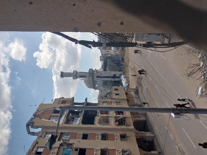 The image depicts a street scene in Khan Yunis, Palestinian Territory, under a partly cloudy sky. A multi-story building on the left exhibits severe structural damage, with large sections of its facade missing, exposing interiors, and numerous bullet holes visible. Blue fabric hangs from a damaged balcony on this building. In the background, a mosque with a dome and a tall minaret is visibly damaged, partially collapsed, and surrounded by extensive rubble. The asphalt road, showing faint lane markings, runs through the center of the frame and is bordered by piles of debris, concrete fragments, and exposed rebar. Several individuals are present on the street: an adult and a child in a green shirt walk in the mid-left, two adults walk together further down the street on the right, and other scattered individuals are seen moving. Overhead utility cables traverse the scene. A streetlight pole stands on the left side of the road. In the immediate right foreground, a partial view of a tan-colored wall with multiple impact marks (bullet holes) is visible.