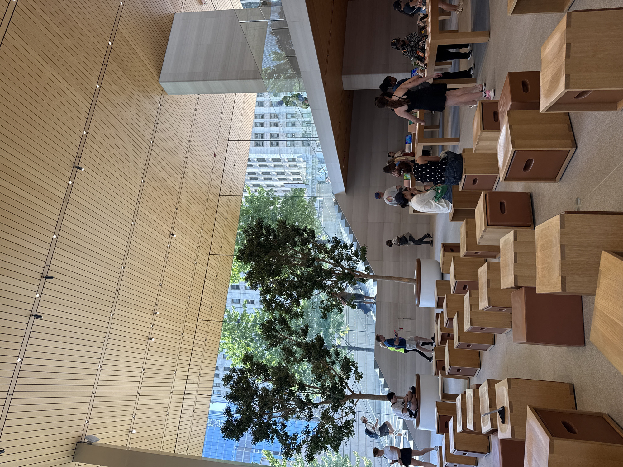 The image depicts the interior of an Apple Store in Chicago, United States of America. The store features a modern, minimalist design with a slatted wooden ceiling and large glass windows offering views of city buildings and trees. In the foreground, numerous wooden cubic seats are arranged on the floor, some with leather accents. Several individuals are seated on these cubes, interacting with electronic devices or conversing. In the mid-ground, a staircase with glass railings leads to an upper level, and two large trees are placed near the stairs, adding a natural element to the space. People are seen walking up and down the stairs and browsing displays of Apple products on wooden tables. The overall environment appears bright, spacious, and contemporary.