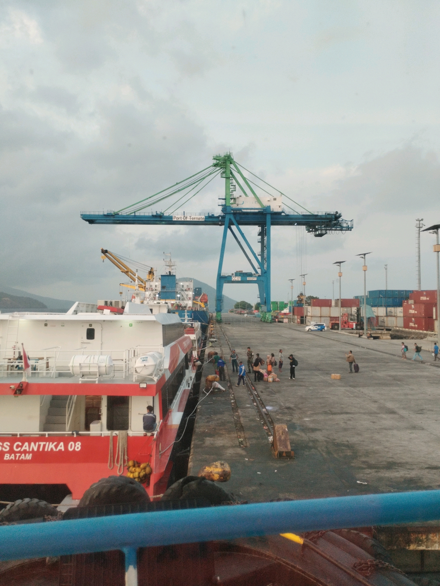 The image depicts a bustling port scene at the Port of Ternate in Ternate, Indonesia. A large cargo ship, identified as "KM. CANTIKA 08 BATAM," is docked at the quay, painted in a striking red and white livery. Several people are visible on the ship's deck and on the dock.
Dominating the skyline is a massive blue and green gantry crane, a symbol of the port's cargo handling operations. The crane has "Port Of Ternate" clearly labeled on its structure. The dock area is paved and shows signs of activity with stacks of shipping containers in the background, some blue and some red.
Several individuals are scattered across the dock, some carrying luggage, suggesting passenger activity or embarking/disembarking operations. A forklift is positioned near the containers, hinting at ongoing cargo movement. The environment appears to be an overcast day, with mountains visible in the distance, suggesting a coastal or island location. The overall impression is one of a busy, operational port with both cargo and passenger elements at play.