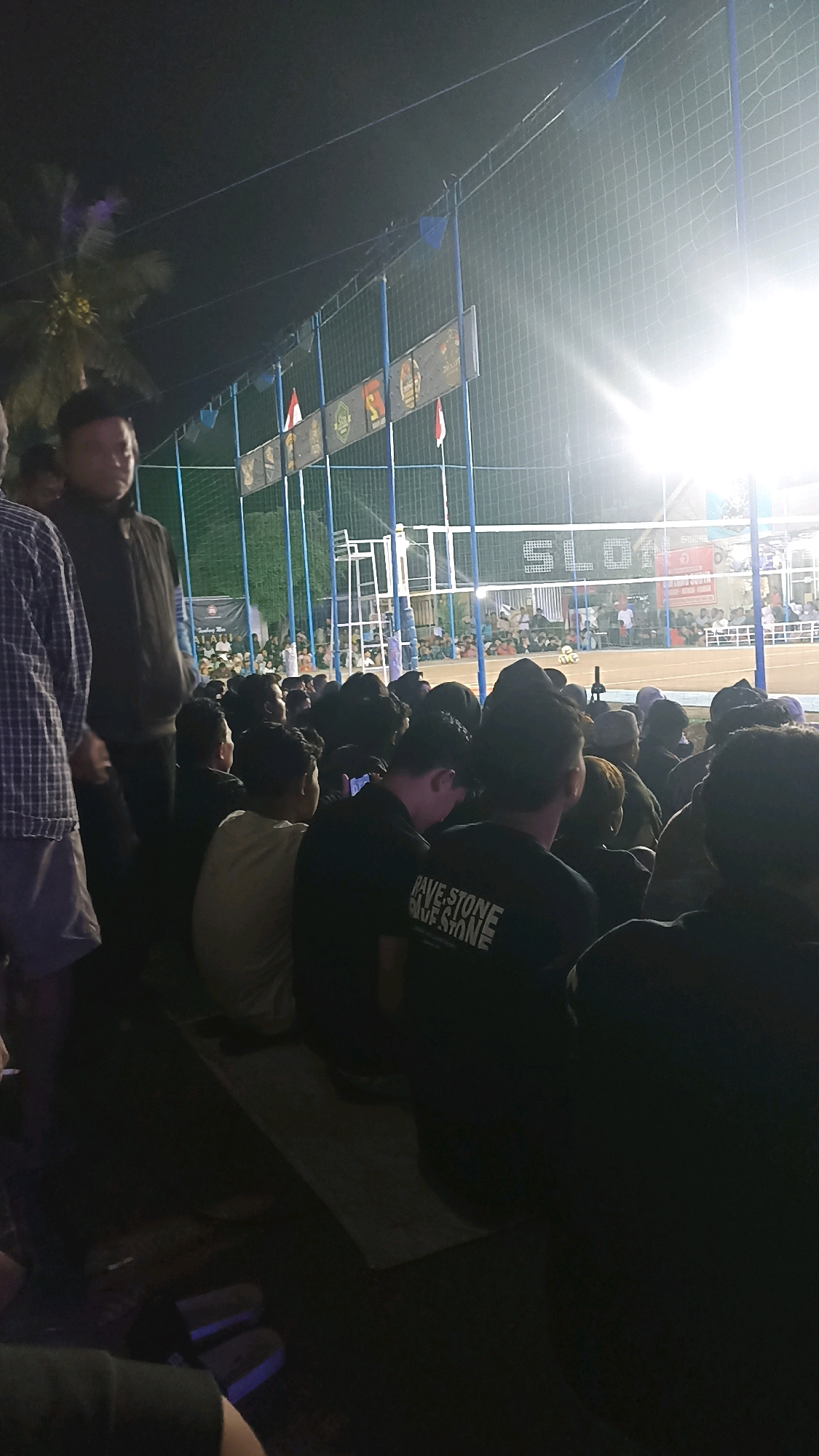 An outdoor volleyball match is underway at night, illuminated by bright lights, with spectators seated on bleachers. The scene is packed with a crowd, many of whom are young men. The court is enclosed by a net and fencing. On the side of the court, banners with various logos and Indonesian flags are displayed. In the foreground, several individuals are visible, some facing the game and others looking down. The image captures a lively community event in Pacitan, Indonesia.