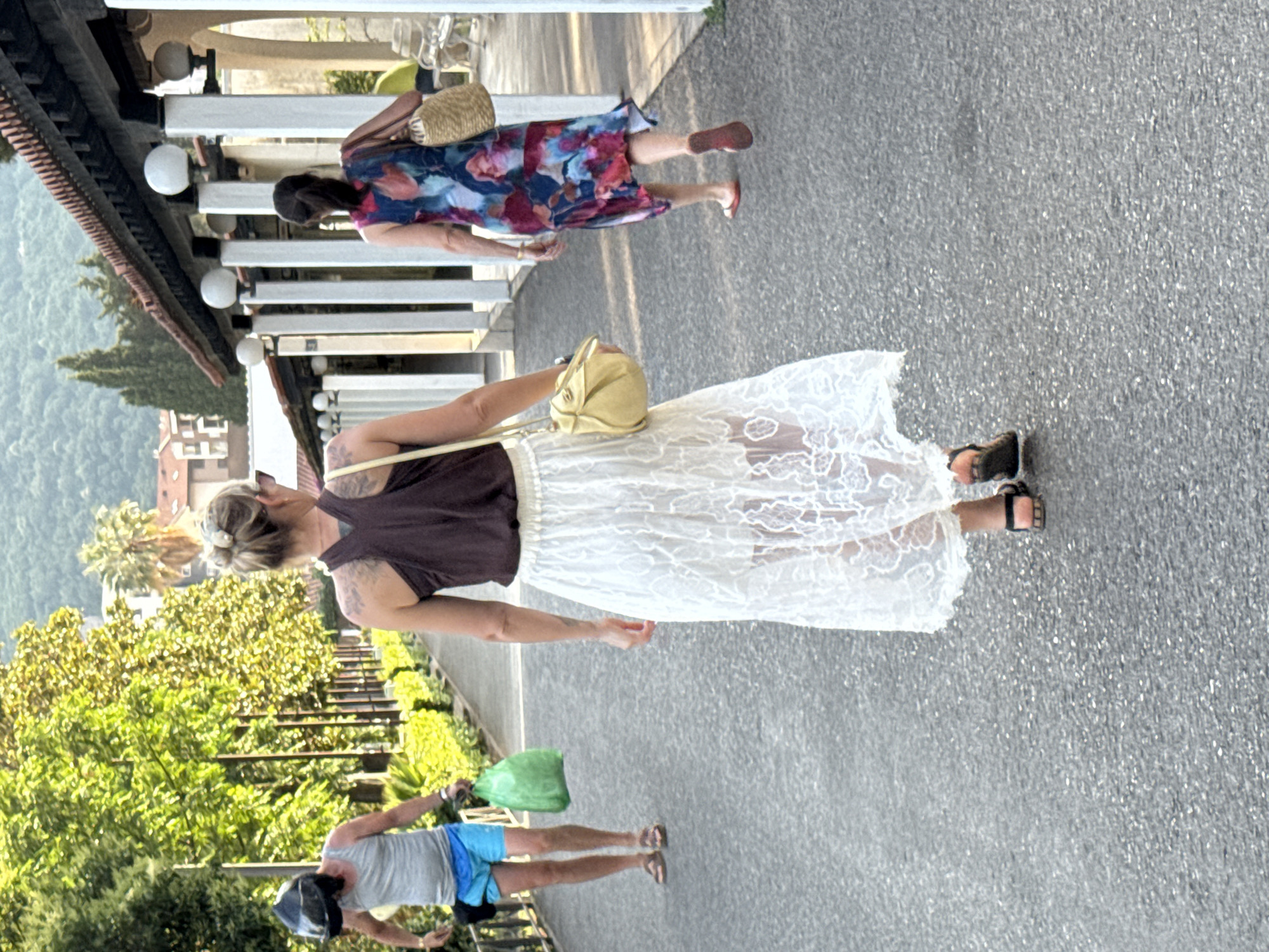 Three individuals are walking away from the camera on a paved walkway lined with trees and a covered walkway with white pillars and globe lights. The person in the foreground, a woman with blonde hair pulled back, wears a dark sleeveless top and a long, white, sheer lace skirt. She carries a light yellow crossbody bag. To her left, further down the walkway, a person wearing a grey tank top, blue shorts, and a motorcycle helmet is walking, holding a bright green plastic bag in their left hand. To the right of the woman in the foreground, another woman walks in a multi-colored floral dress and carries a woven straw bag over her right shoulder. The background shows buildings with red tiled roofs and green foliage, with a mountainous landscape visible in the distance. The scene appears to be in an outdoor public area, possibly a park or a promenade, with architectural elements suggesting it could be within a resort or a historical complex. Based on the architecture and natural environment, the location is identified as the promenade in front of the Avala Resort and Villas in Budva, Montenegro.