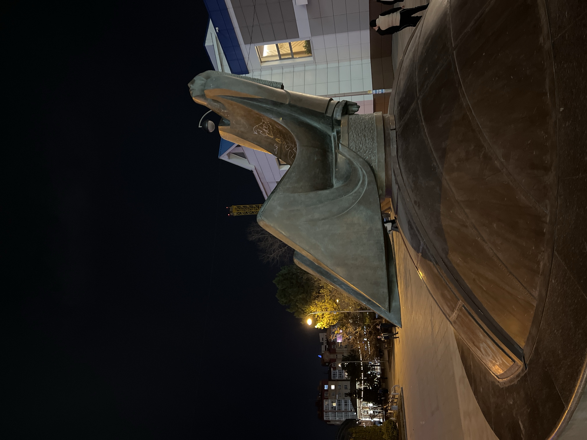 The image shows a night view of the "Globe" sculpture in Tyumen, Russia. The bronze sculpture dominates the right side of the frame. In the background is a building with lit windows. Further back, illuminated streetlights line a street with multi-story buildings on either side. The rest of the view is black, suggesting night. Two people can be seen in the upper right of the frame, partially out of view.