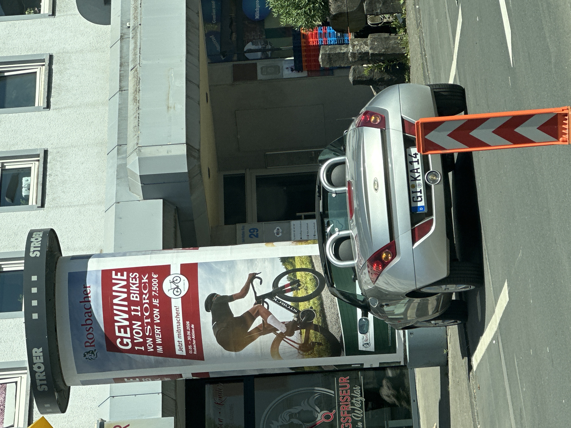 The image depicts an outdoor scene in Dillfeld, Germany, featuring a parked silver Ford Focus convertible with a license plate "GI KA14." A large Stroer advertising pillar stands alongside the vehicle, displaying an advertisement for Rosbacher, promoting a contest to win one of 11 Storck bicycles. The ad includes a cyclist graphic and promotional text. A temporary chevron patterned traffic barrier is positioned directly behind the car. Behind the car and advertising pillar is a commercial building with visible businesses: a hairdresser and a business selling healthy goods.