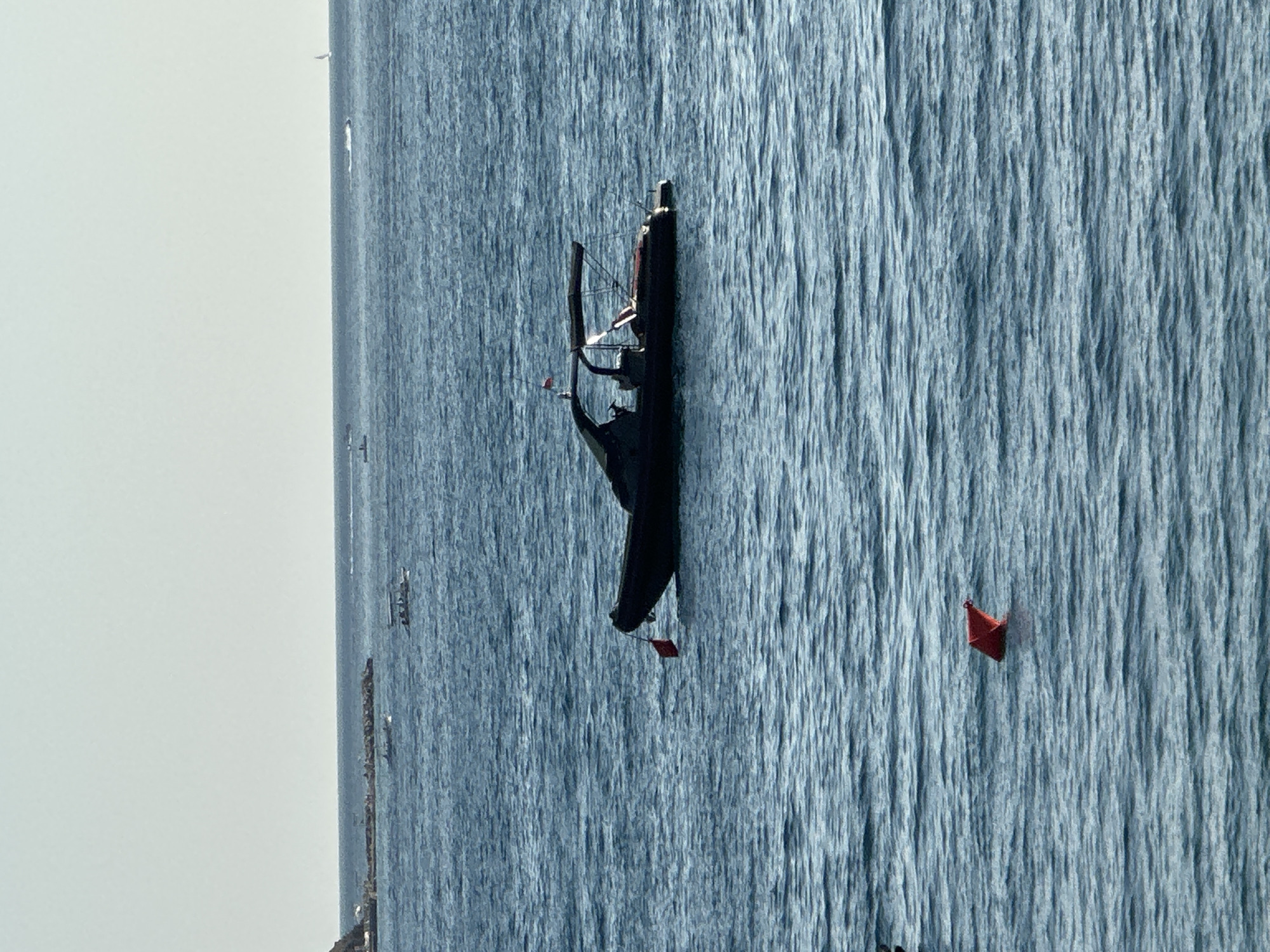 A high-angle, medium shot captures the Adriatic Sea off the coast of Budva, Montenegro. The water is a deep, rippling blue under a clear, pale sky. In the mid-ground, a dark-hulled RIB (Rigid Inflatable Boat) with a canopy is anchored, a small red flag fluttering from its bow. To the left and slightly forward of the RIB, a smaller red conical buoy bobs on the surface. In the far background, a breakwater lined with rocks extends from a distant shoreline, with several small boats visible near it. The overall scene is serene and nautical.