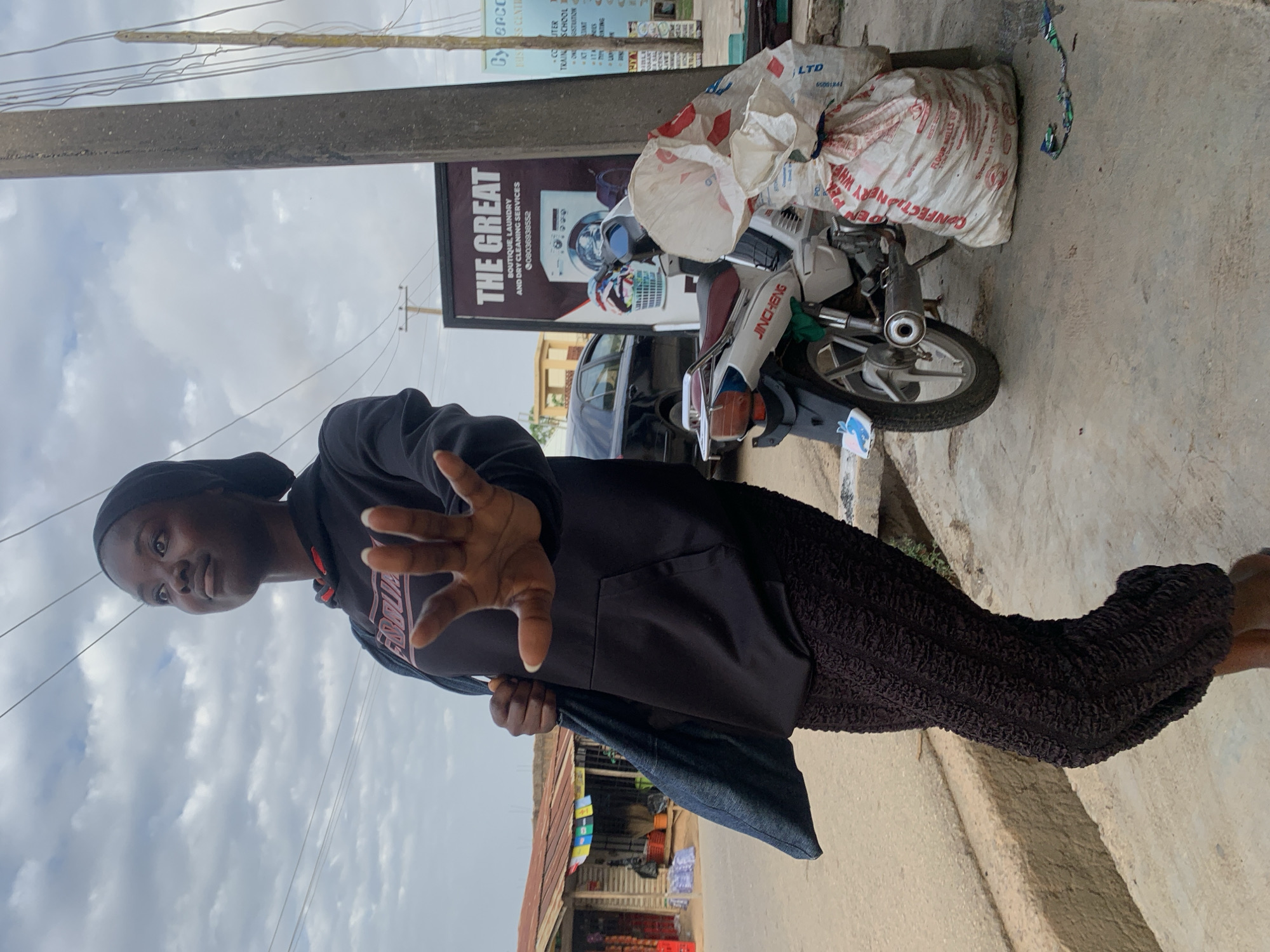 A person with dark hair and a dark-colored hoodie stands in the foreground, their hand extended towards the camera. Behind them is a JINCHENG motorcycle loaded with large white sacks. A sign for "THE GREAT Boutique, Laundry and Dry Cleaning Services" is visible next to the motorcycle. In the background, a storefront with a variety of goods is present. Power lines stretch across the sky. The presence of local businesses like "Cyperco Business Center" and the laundry service, along with the style of architecture and goods displayed, suggest a location in Jos, Nigeria.