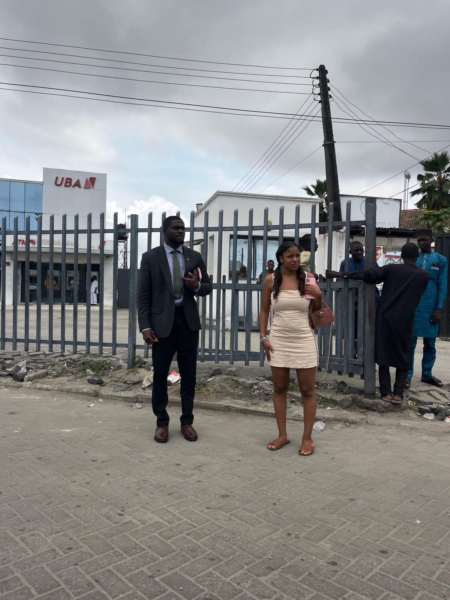 In Ado, Nigeria, in front of a United Bank for Africa (UBA) building, a man in a dark suit, white shirt, and green tie stands to the left. He is looking to his right, with his right hand holding a red object and his left hand at his side. To his right, a young woman in a short, cream-colored dress and flat sandals stands holding a pink phone and a brown purse. Behind them, a gray metal fence encloses the bank. Several other individuals are visible in the background, some standing near the fence and others inside the bank, where ATMs are visible. The ground in the foreground is paved with interlocking bricks and littered with debris. Utility poles and wires are visible against an overcast sky.