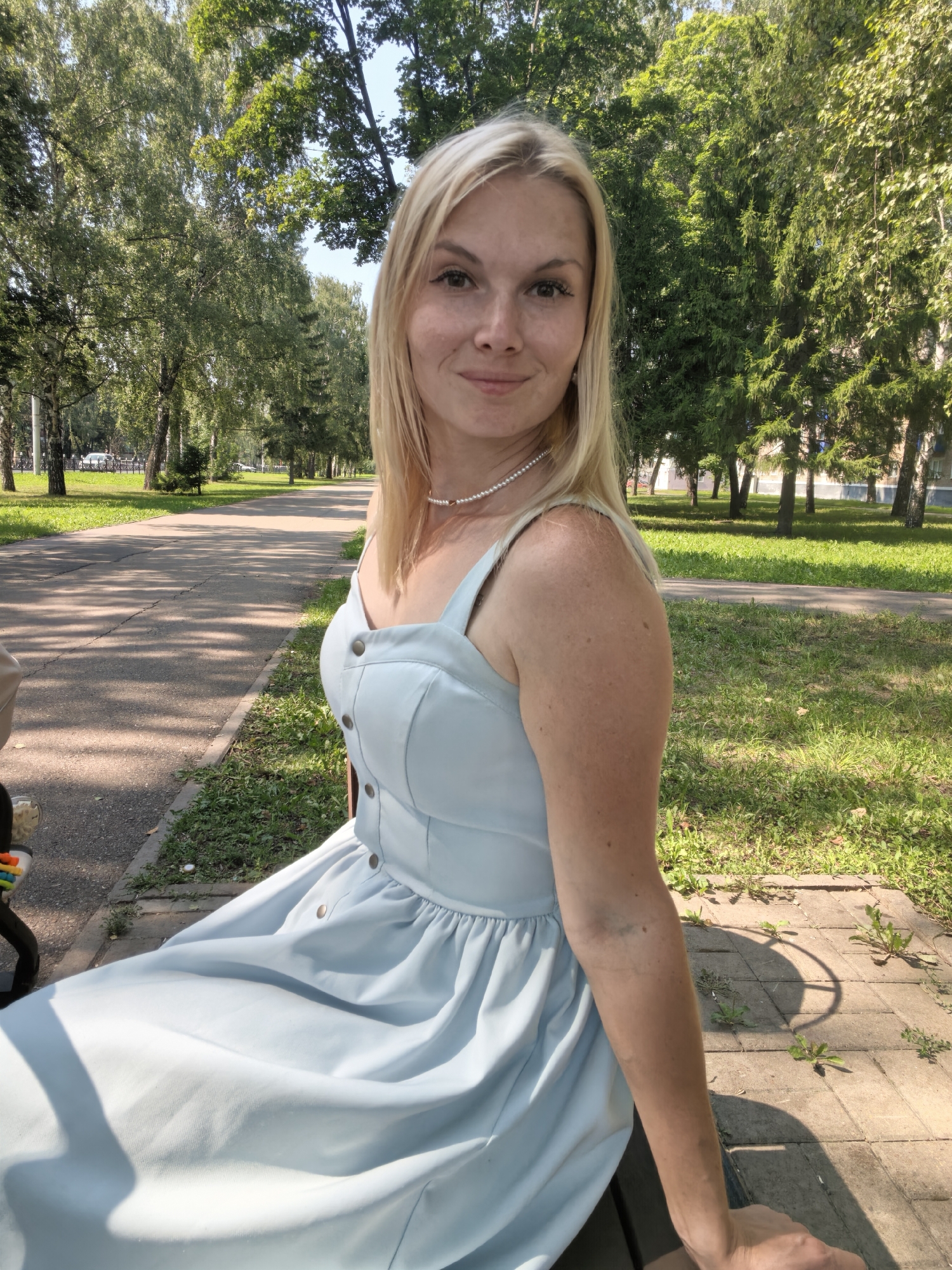 A blonde woman with fair skin and freckles is sitting on a park bench. She is wearing a light blue sleeveless dress with buttons down the front and a pearl necklace. Her hair is shoulder-length and parted to the side. She is smiling at the camera. The park appears to be a tree-lined path with a paved walkway. There are trees and grass visible in the background, as well as buildings in the distance. The lighting suggests it is daytime. The woman is in the city of Sterlitamak, Russia.