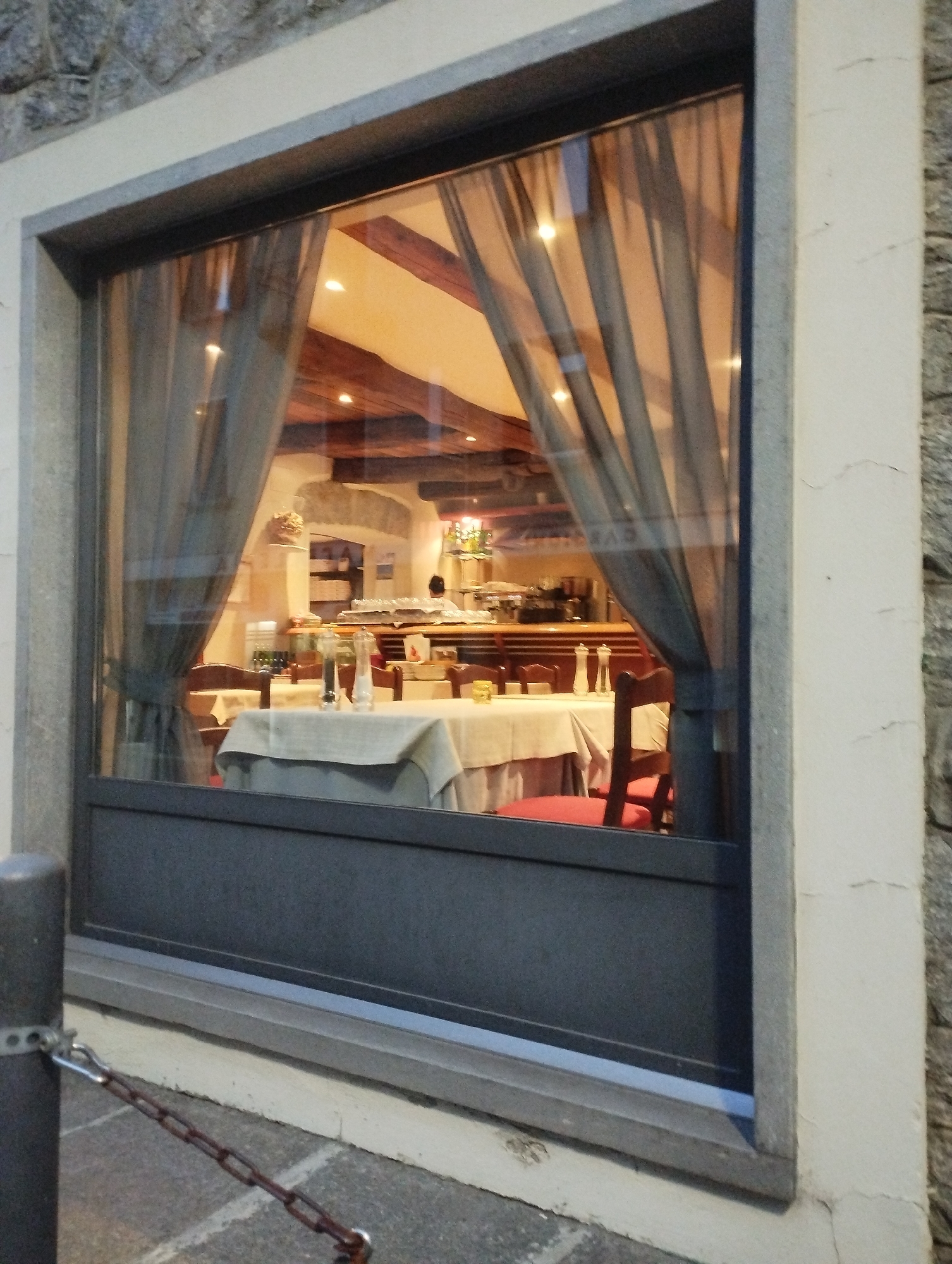 A view through a window reveals the interior of a restaurant. The room features wooden beams on the ceiling and is furnished with tables covered in white tablecloths. Red chairs are visible around some of the tables. In the background, a person is seen behind a counter, and shelves with items are present. Curtains hang on either side of the window, partially obscuring the view. Outside the window, a chain is attached to a metal post, and cobblestone pavement is visible. The building exterior is made of stone and a light-colored stucco.
