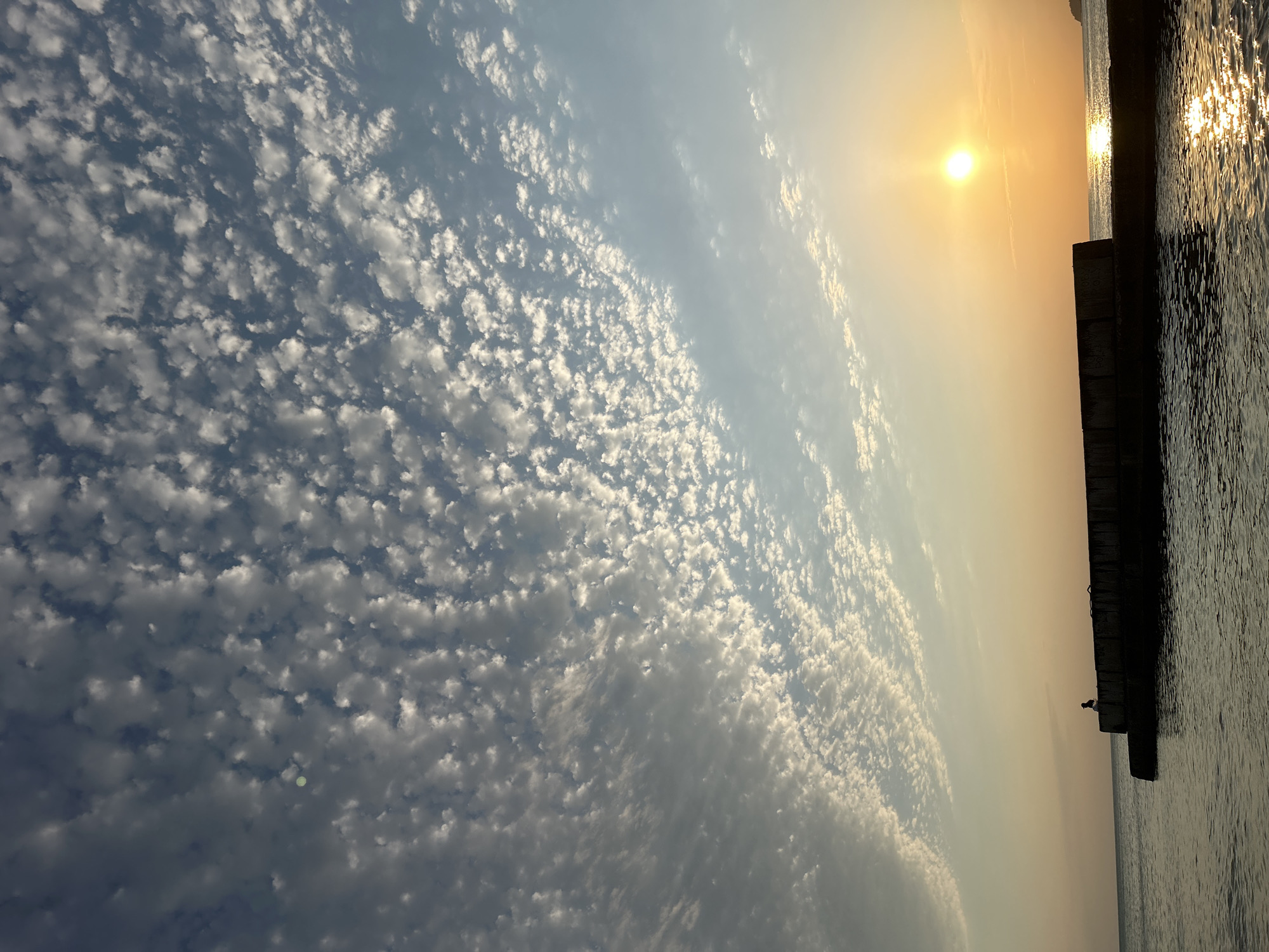 A solitary figure, silhouetted against the setting sun, sits on the edge of a concrete breakwater. The sky above is a dramatic canvas of clouds, with small, scattered white formations creating a textured pattern against a gradient of blue and pale orange. The sun, a bright orb, casts a shimmering golden path across the calm water. The water itself appears dark and reflective, with gentle ripples. The breakwater is long and dark, with a rough texture. The figure is positioned on the left side of the breakwater, facing towards the water. The overall scene suggests a peaceful, contemplative moment at dusk, likely by the sea. The location is Dzeberkoi, Russia.