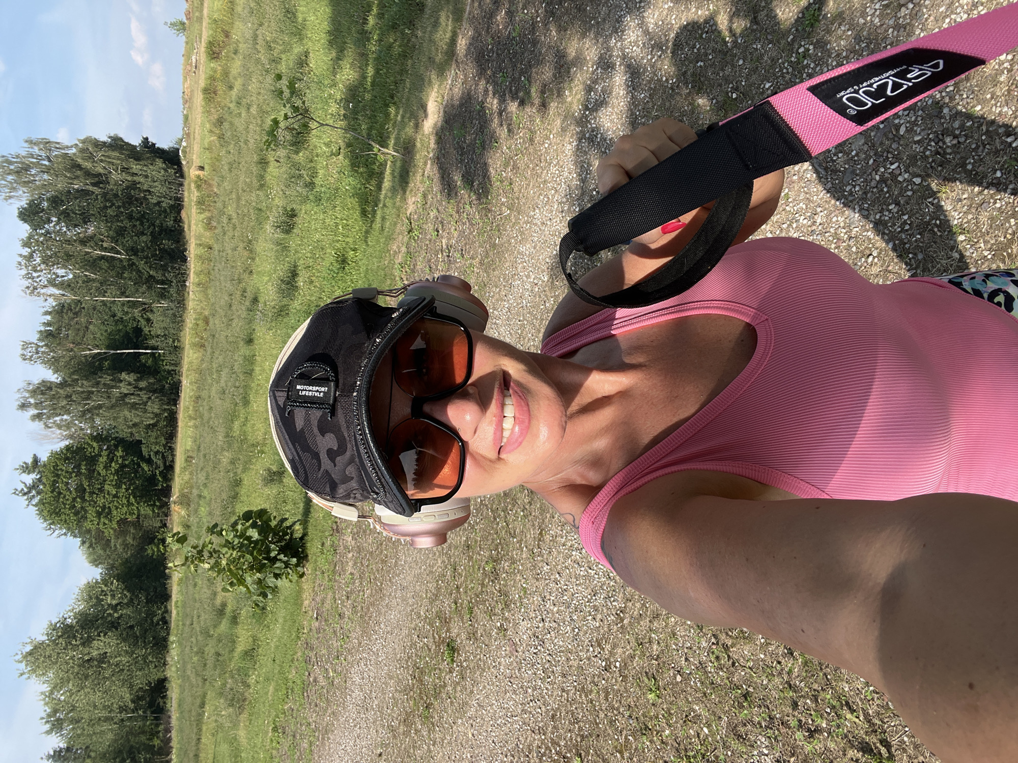 The image depicts a woman taking a selfie outdoors on a sunny day in Hodyszewo, Poland. She is wearing a pink tank top, a camouflage baseball cap, and large sunglasses. Over-ear headphones are also visible. In her right hand, she holds a black and pink strap with the logo "ARIZJO" and text indicating "PHYSIOTHERAPY & SPORT." The background features a gravel path, a grassy field with scattered trees, and a forest line under a blue sky with scattered clouds. The overall impression is that of a person engaged in outdoor activity or exercise.
