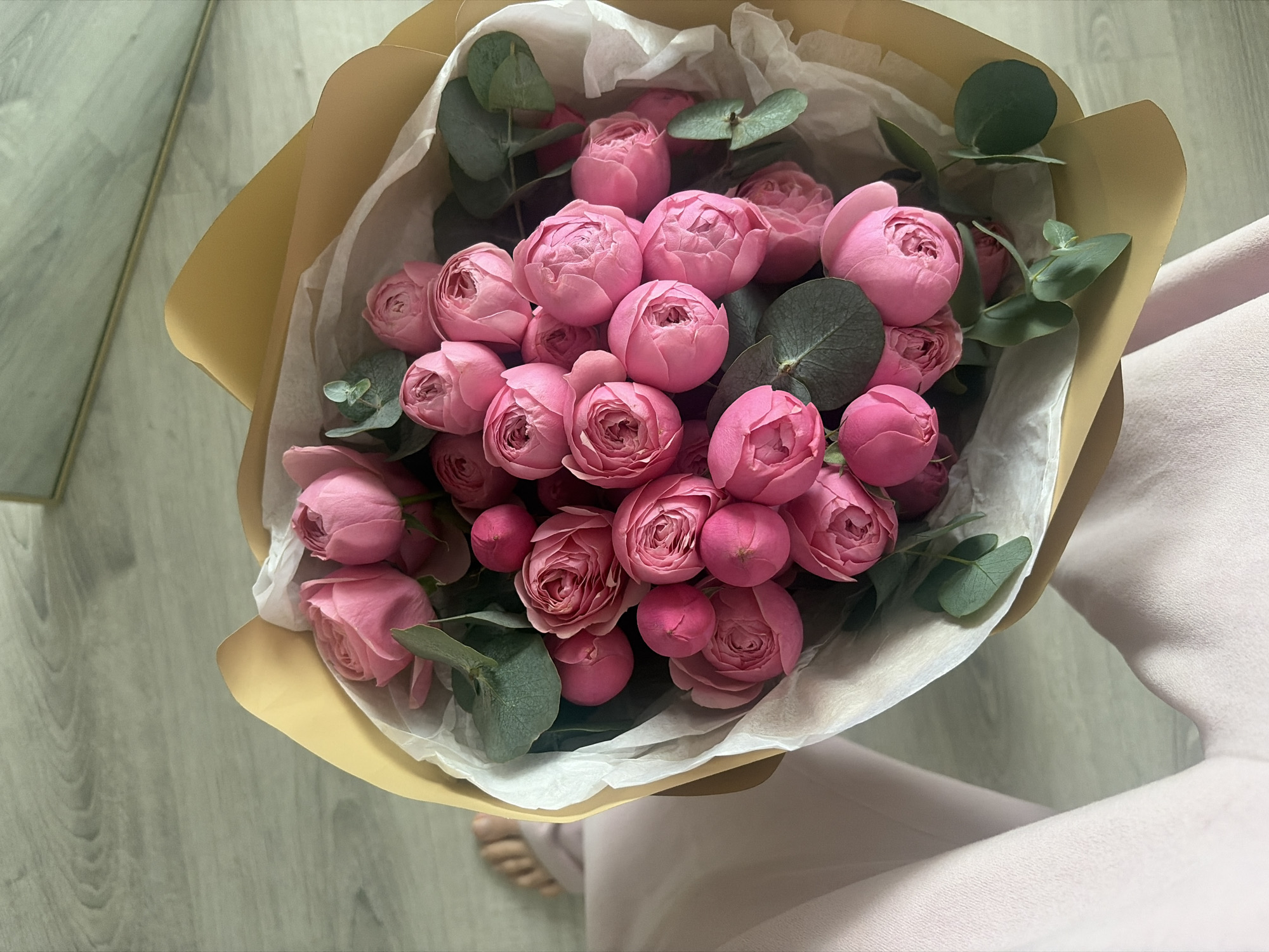A top-down, medium shot captures a bouquet of pink roses and eucalyptus leaves. The flowers are tightly clustered, with some buds still closed and others beginning to unfurl their delicate petals. The eucalyptus leaves, a deep green, provide a contrasting texture and color to the soft pink of the roses. The bouquet is wrapped in thin, white paper and a sheet of light brown paper. The wrapper extends from the top of the image, with the flowers and leaves spilling out from its center.
In the background, a light wood-grain floor is visible. To the left, a reflective surface, possibly a mirror or a window, shows a muted green, textured pattern. To the right, a person's lap is visible, covered in a soft, light pink fabric that suggests clothing or a tablecloth. The person's bare feet, with visible toes and toenails, are also visible at the bottom center of the frame, resting on the wooden floor. The lighting is soft and diffused, creating a gentle atmosphere. There is no visible text in the image. The overall impression is one of simple elegance and natural beauty, perhaps a gift or a decorative arrangement.