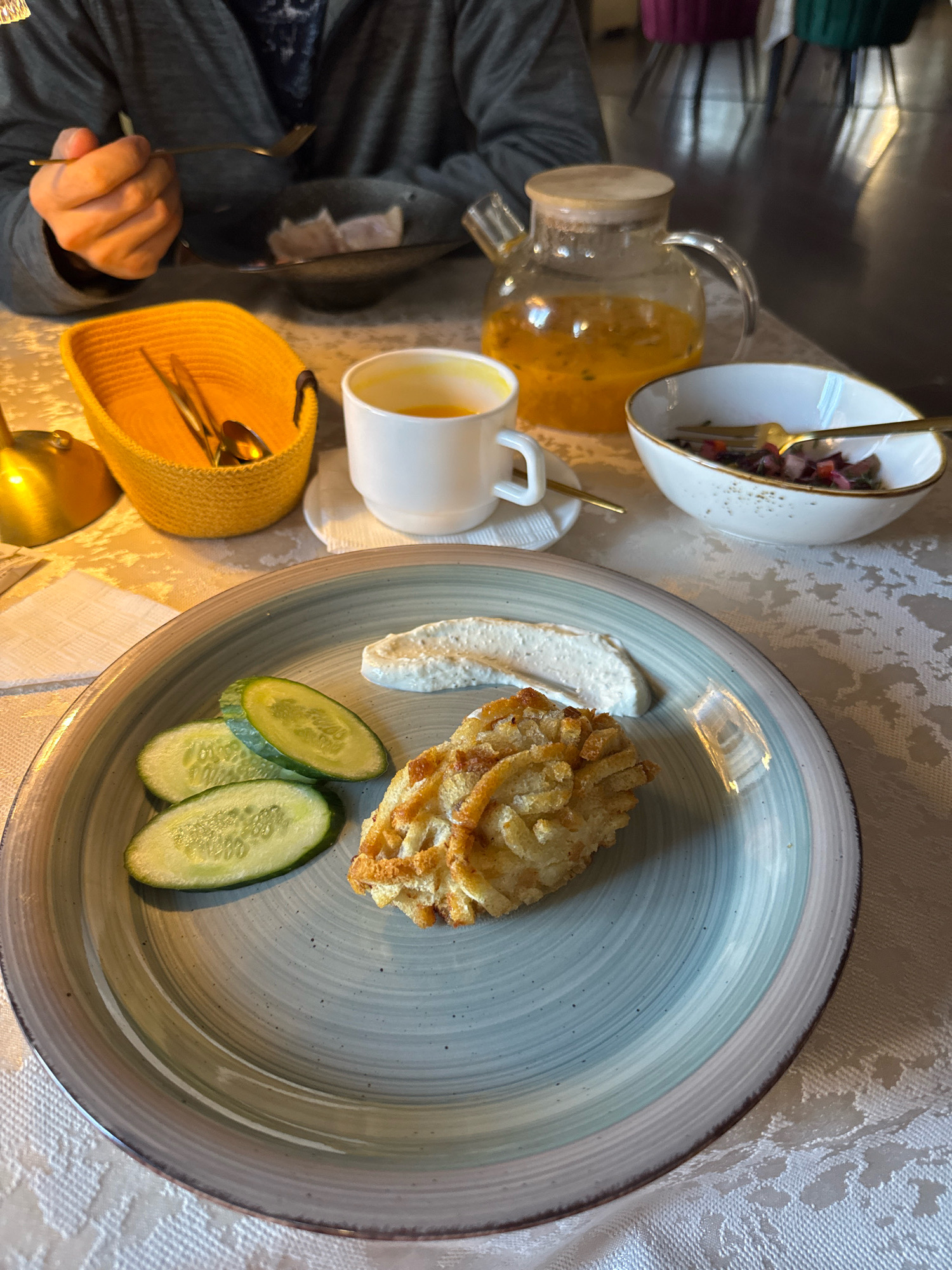 A close-up shot shows a table setting with food and drinks, potentially at a restaurant in Torzhok, Russia. In the foreground, a grey-blue plate holds a meal consisting of three cucumber slices, a dollop of white spread, and a golden-brown fried item. To the left, a yellow woven basket contains cutlery. Behind it, a white teacup with an orange liquid sits on a saucer. A glass teapot filled with a similar orange liquid is positioned further back, next to a white bowl containing a salad with red and green elements and a fork. In the background, a person's hands are visible, one holding a fork. The table is covered with a white tablecloth featuring a textured pattern. The scene suggests a meal is being served or enjoyed.