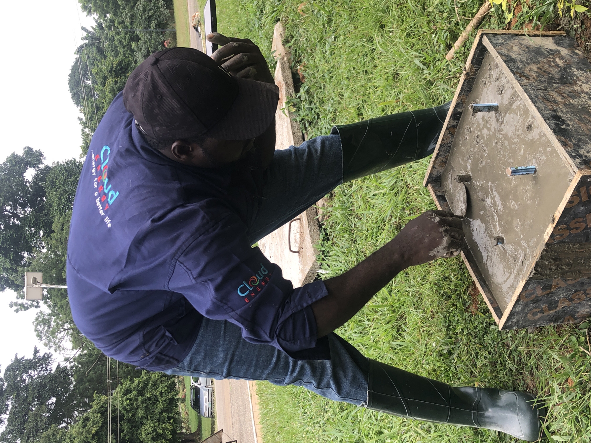 A Nigerian man, wearing a dark blue long-sleeved shirt with a "Cloud Energy" logo and blue jeans, and black rubber boots, is performing concrete work outdoors. He is bending over a square wooden mold filled with wet concrete, using a trowel to smooth the surface. Two threaded metal rods are partially submerged in the concrete. The background features lush green grass, trees, and a paved road with a silver car. A solar-powered street light is visible in the distance. The scene is likely located in Iju, Nigeria, a town known for its waterworks and power generation facilities.
