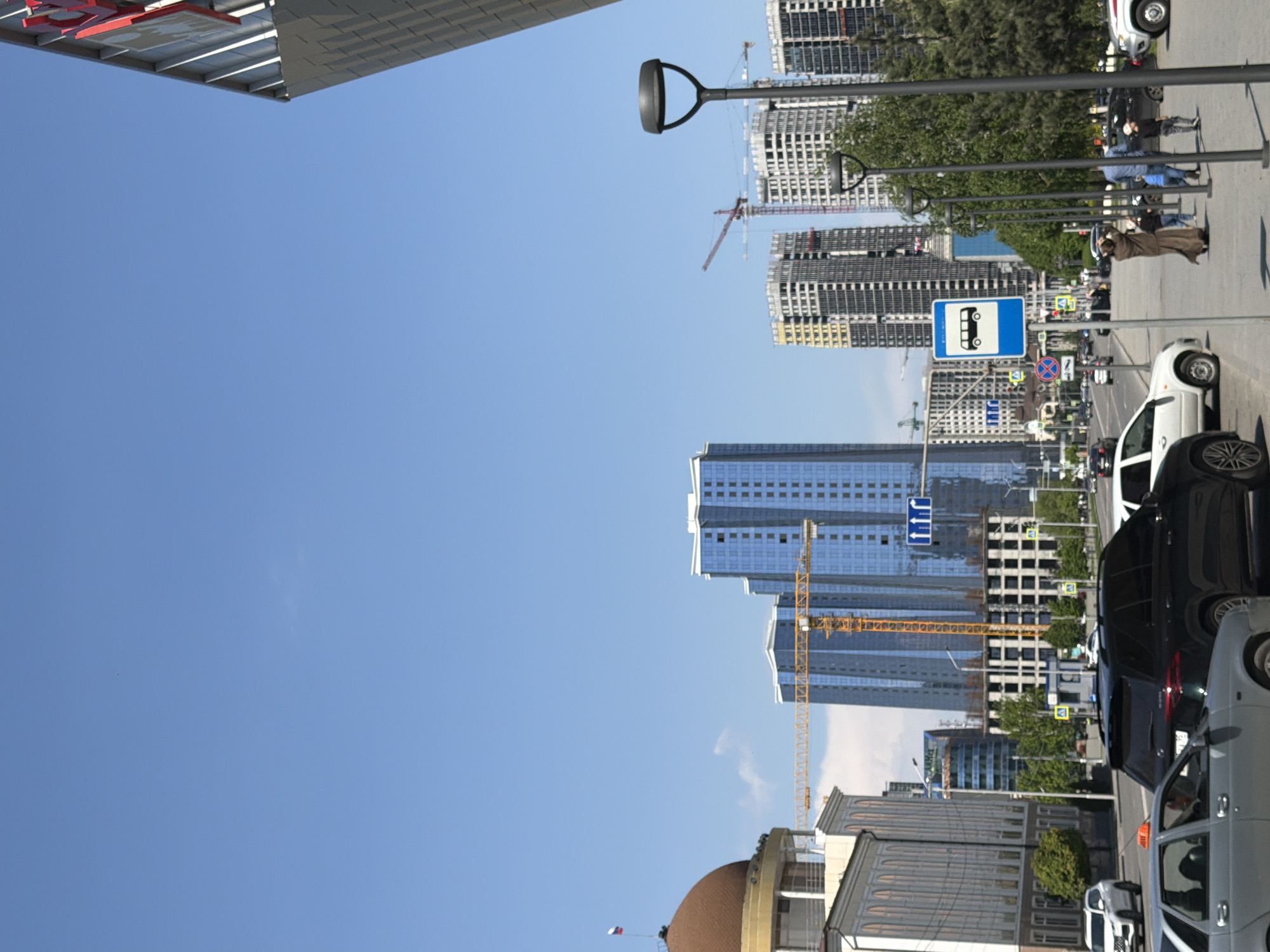 The image captures a street scene in Grozny, Russia, under a clear blue sky. Numerous modern high-rise buildings are present, some under construction, indicated by construction cranes. A row of streetlights line the road, alongside pedestrian traffic and parked cars. A bus stop sign is visible. Pedestrians are walking on the sidewalk. Traffic is present on the road. Buildings display a modern architectural style.