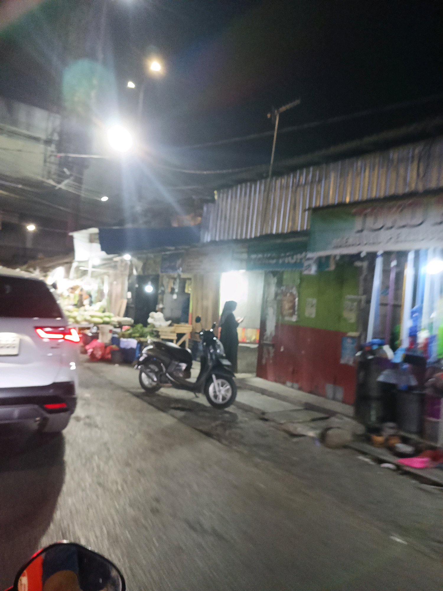 The image captures a street scene at night in what is identified as Makassar, Indonesia. A white SUV is parked on the left side of the road, facing away from the camera. In front of the SUV, a dark-colored scooter is parked, with its front wheel turned slightly to the left. A person wearing a hijab, dressed in dark clothing, stands next to the scooter, facing towards the right.
The background features a row of small businesses or market stalls. These structures have corrugated metal siding and some have awnings or tarps. Some stalls appear to be selling produce, with stacks of green items visible. Lighting is provided by overhead lamps, casting a dim glow on the scene. The road surface is asphalt and appears wet, possibly from rain. The overall impression is of a busy, modest marketplace operating at night.
