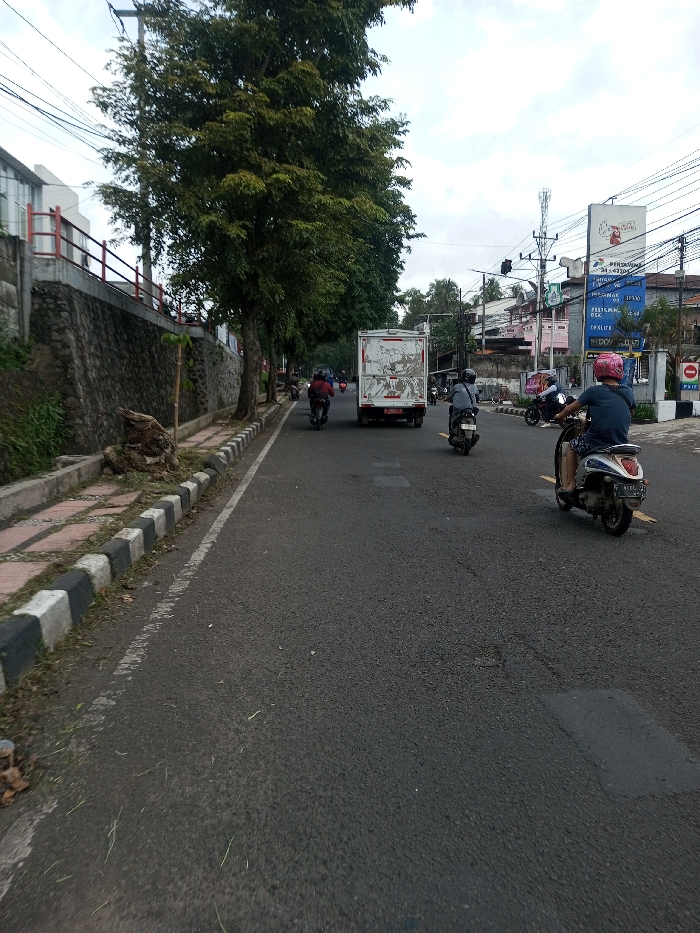The scene depicts a street in the city of Khajuripada, India, under an overcast sky. The asphalt road, showing patches and white lane markings, occupies the central and right portions of the frame. To the left, a concrete sidewalk with black and white checkered curbing borders a stone retaining wall. A line of mature trees with green foliage stands along the left side, behind the sidewalk and wall.
Multiple vehicles are in motion, traveling away from the camera. In the foreground right, an individual wearing a blue shirt, shorts, and a pink helmet operates a light-colored scooter with a visible license plate number "4279." To the left and slightly ahead, another person on a dark scooter, wearing a grey helmet and dark attire, proceeds. Further into the scene, a white box truck with a red bumper and partially obscured rear license plate (beginning with 'B') is positioned in the left lane. Behind the truck, two additional individuals on dark motorcycles are visible, one wearing a red jacket.
The background on the right side reveals a commercial urban environment featuring multiple buildings, utility poles, overhead power lines, and a cellular tower. Prominent signage includes a large white billboard displaying text such as "PESONA," "METRO," "15.000," "34 - 36," and "DO NOT LOAD." A red circular 'no entry' sign is also visible. A traffic light assembly is present above the distant intersection.