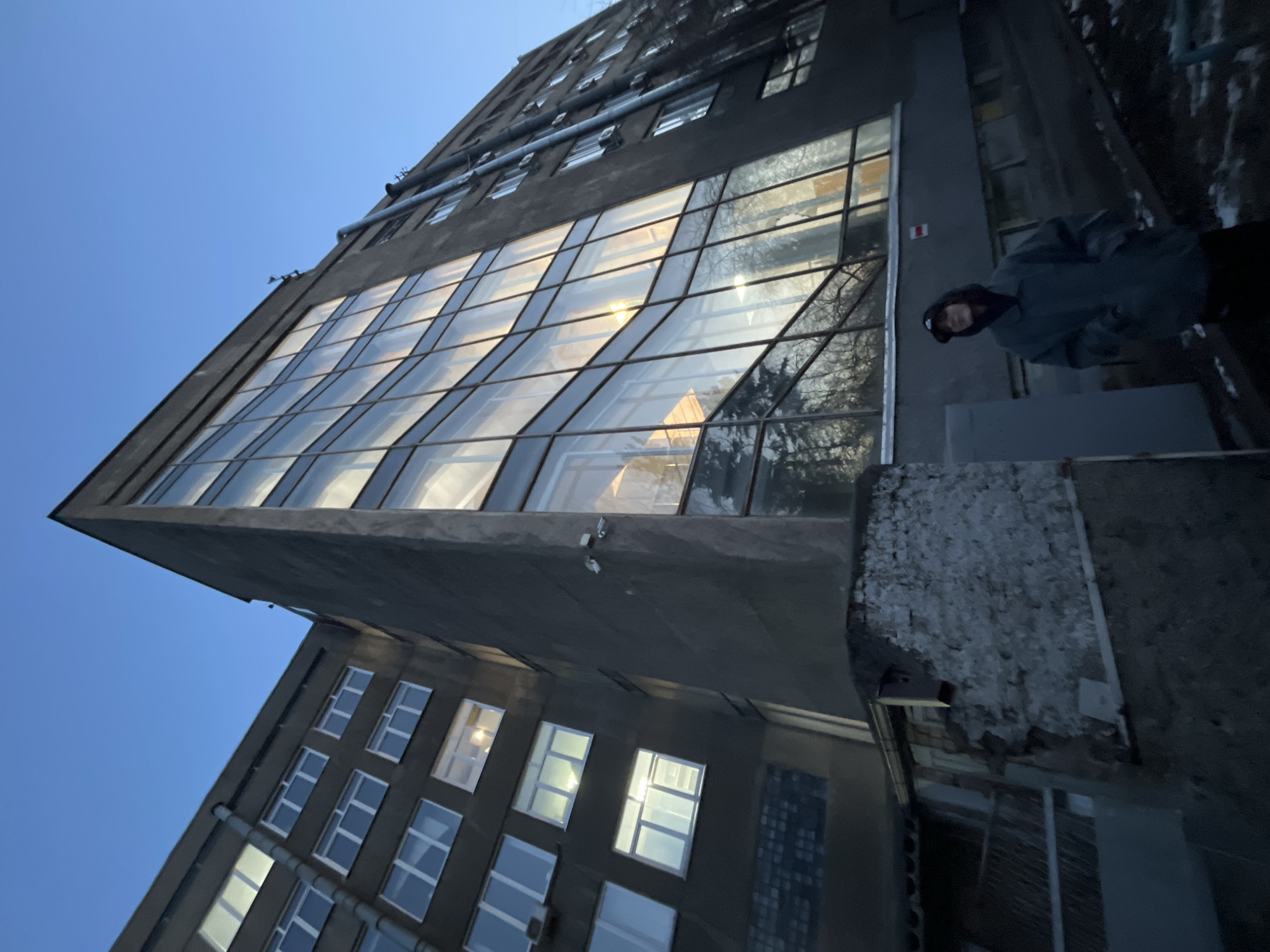 A low-angle, wide shot captures a tall, angular building under a clear blue sky, presenting a modern, somewhat industrial aesthetic. The main structure features a prominent, multi-story glass facade that slants upwards and reflects warm, golden light and the bare branches of trees, suggesting either sunrise or sunset. Above and to the left of this glass section, the building's concrete exterior is dark gray, adorned with several horizontal pipes and rows of smaller, square windows.
On the right side of the image, a person is visible, standing on a dark, textured ledge or walkway adjacent to the building. They are dressed in a dark hooded jacket or coat and a dark beanie, looking towards the camera with a subtle expression. The concrete ledge beneath them shows patches of white, possibly snow or frost, hinting at cold weather conditions.
To the lower left, another section of the building, or a connected structure, is visible, also with a dark gray facade and numerous smaller, rectangular windows, some of which are illuminated from within, casting a soft white glow. The overall lighting is dim on the concrete surfaces but bright in the sky and reflections, emphasizing the stark contrast of the architecture against the clear sky. No discernible text is visible.