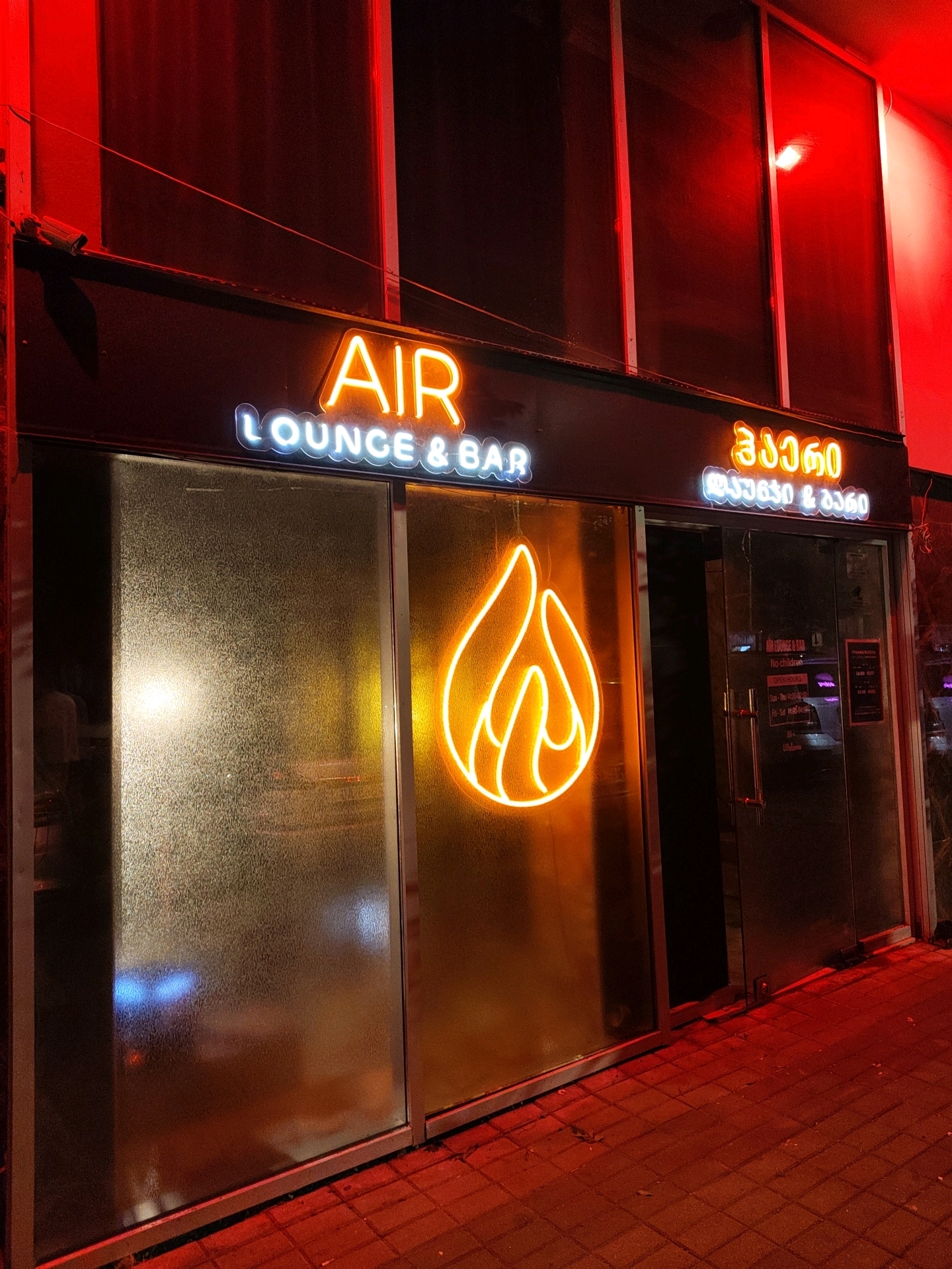The image shows the exterior of a lounge and bar named "AIR" located in Batumi, Georgia. The establishment features prominent neon signage, with "AIR" in orange letters and "LOUNGE & BAR" in white letters. A large orange neon flame logo is also displayed on the frosted glass facade. To the right of the entrance, there is Georgian text in neon, likely indicating the name of the establishment or a slogan. The building has large glass windows and a dark doorway. The scene is illuminated by red ambient light and the glow of the neon signs. The ground outside appears to be paved with brick-like tiles, reflecting the red light. No people are clearly visible in the immediate vicinity of the entrance.