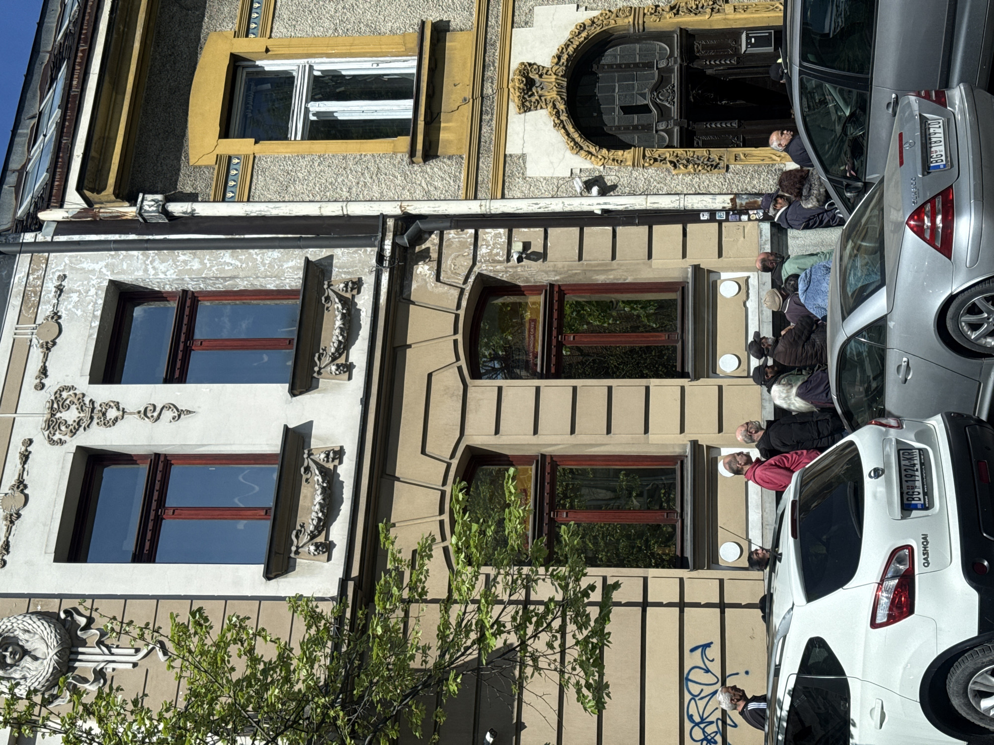 The image depicts a street scene in Belgrade, Serbia. Two buildings with distinct architectural styles are visible. The first building has a white facade with ornate carvings and two windows with red frames. The second building has a beige facade with simpler detailing and two windows with red frames and one ornate doorway. People are sitting on the steps in front of the second building, near a silver and a white car that are parked on the street. The license plate number of the silver car is BG 1879TO and the license plate of the white car is BG 1924KR. There is graffiti visible on the wall of the second building near the white car. A small tree is in the foreground. The image is rotated 90 degrees counter-clockwise.