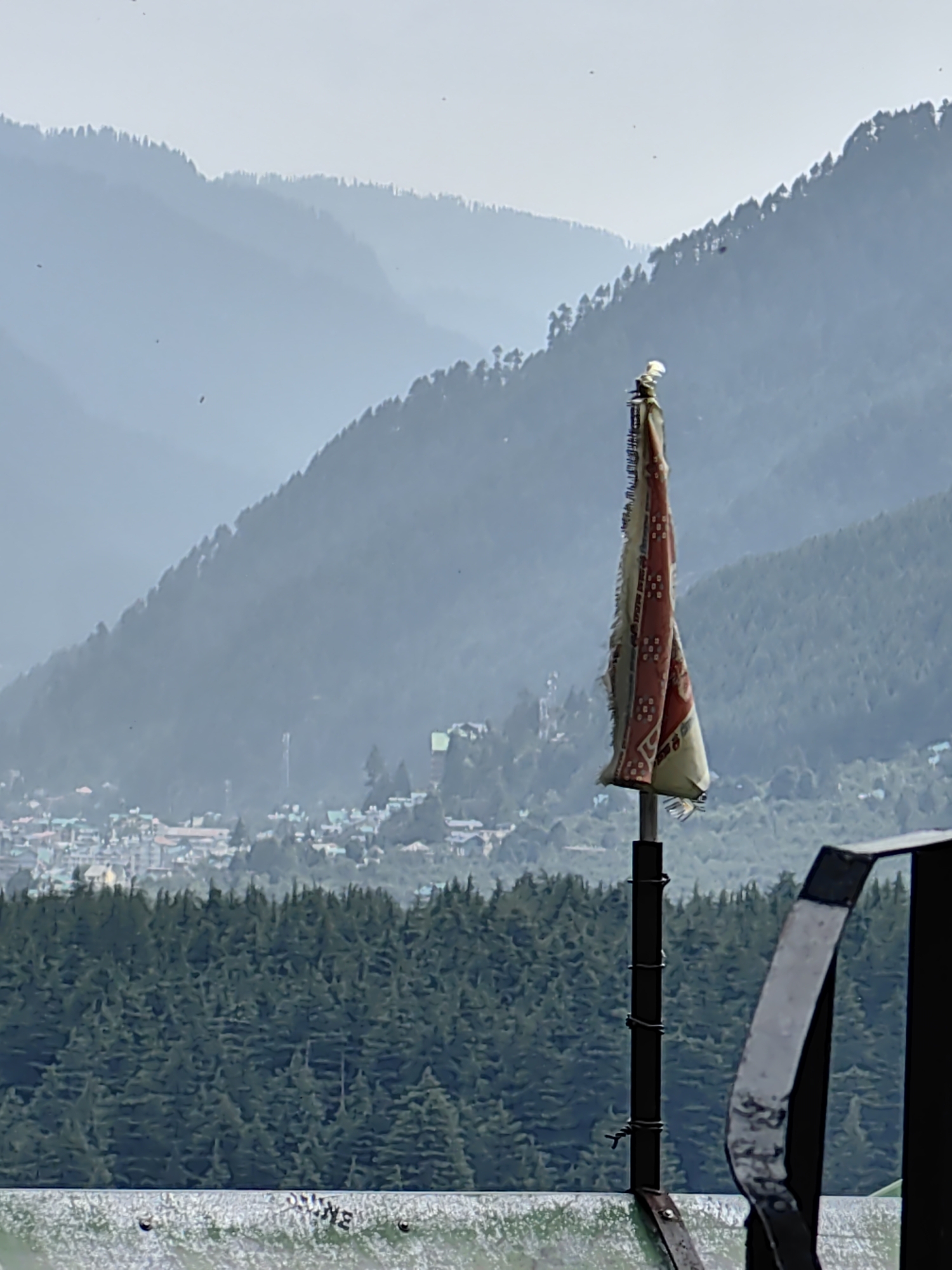 The image depicts a mountainous landscape with a village nestled in the valley. In the foreground, a tattered flag on a pole is visible, attached with wire. Beyond the flag, a dense forest of pine trees covers the lower slopes of the mountains. A town with buildings of various colors, many with green roofs, is situated in the middle ground. The background shows layered mountains fading into a hazy sky. The overall scene suggests a remote, high-altitude location, consistent with the town of Vashisht in India, known for its hot springs and proximity to the Himalayas.