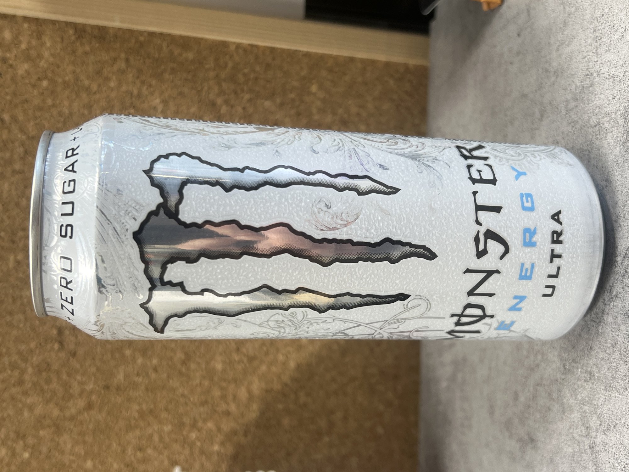 A white Monster Ultra energy drink can sits on a gray surface. The can is oriented horizontally. Text on the can reads "ZERO SUGAR +" and "MONSTER ENERGY ULTRA." The background consists of a tan wall on the left side and the gray surface on the right. There are no people in the image. The location cannot be determined to be Sofia, Bulgaria.