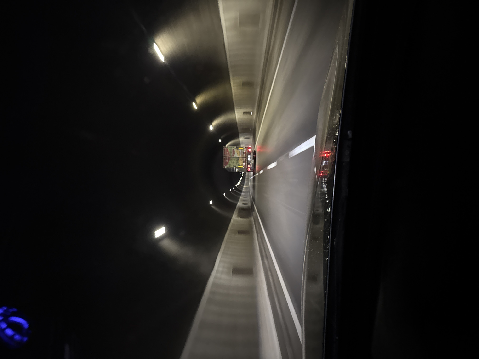 The image shows the interior of a tunnel, likely a road tunnel, in Dolna Vasilitsa, Bulgaria. The tunnel is dark, illuminated by a series of evenly spaced lights along the tunnel's curved ceiling. A truck with visible red taillights is seen further inside the tunnel. The viewer is situated within a vehicle, as a window frame is visible on the right side of the image, with what appears to be rain droplets on the glass. The perspective is slightly skewed, giving an off-center view of the tunnel's roadway and walls.