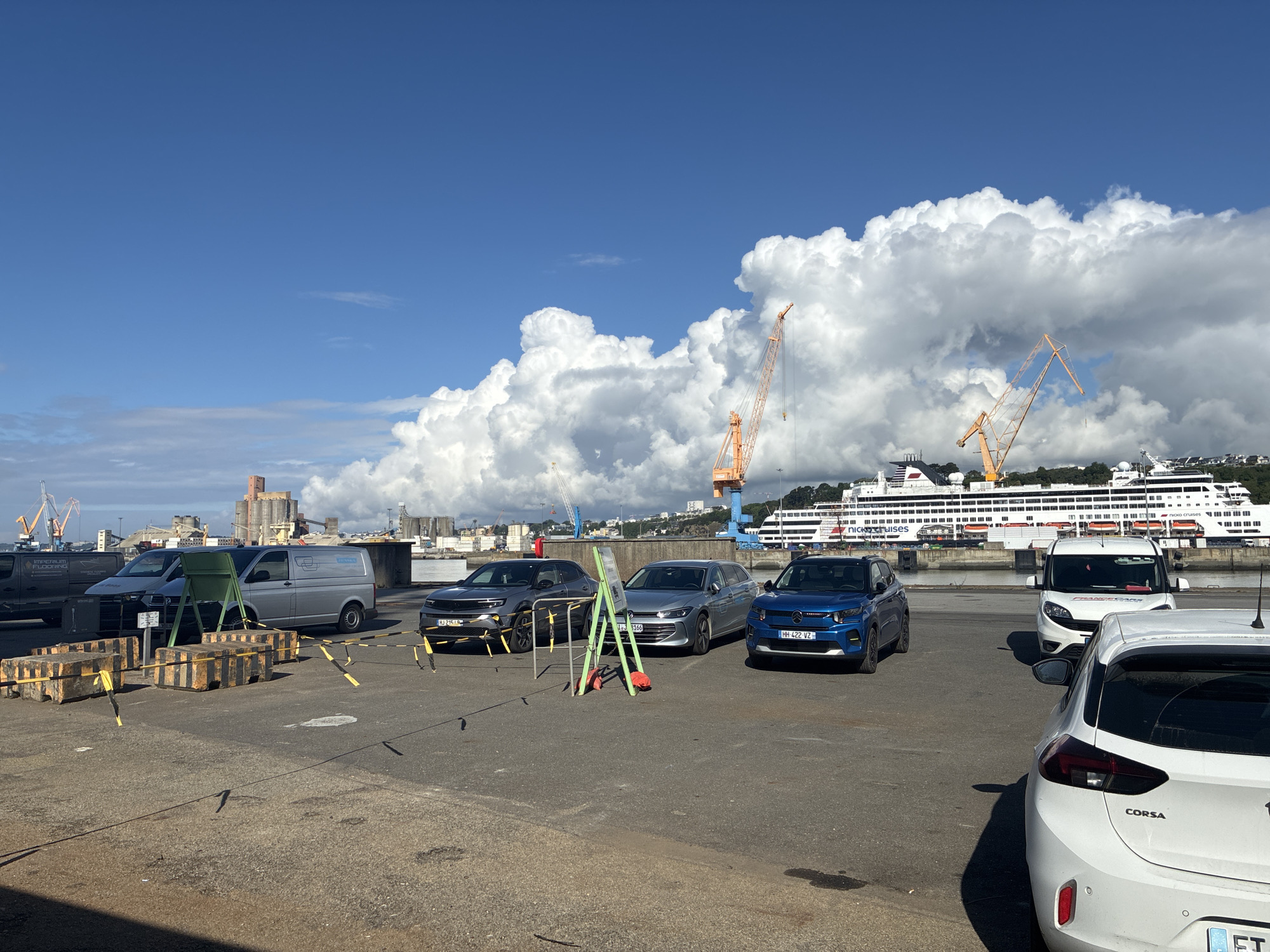 The photograph depicts a parking area in Brest, France, situated near a waterfront, with several vehicles and industrial elements in view. The sky is partially covered with large, cumulus clouds. In the background, a large cruise ship, identified as "nicko cruises," is docked near orange harbor cranes. Vehicles in the foreground include a dark gray SUV with license plate HJ-215-IS, a silver station wagon, license plate 1J8166, a blue compact SUV, license plate HH-422-VZ, a white van, and a white Opel Corsa. Green and red metal A-frame barriers are positioned next to the silver car. Yellow and black striped concrete barriers and caution tape mark off a section of the parking area. To the left, a silver Volkswagen van with the word "Jenneau" on its side and a smaller black van are partially visible. The environment appears to be an industrial port or harbor, with additional buildings and structures in the distance.