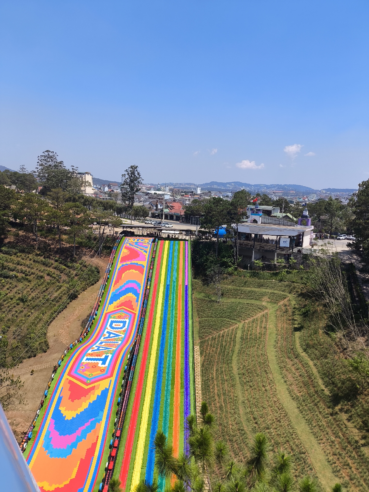 The image captures a scene in Da Lat, Vietnam, specifically featuring a large, colorful slide, likely a tourist attraction, adorned with the word "DALAT" in large, stylized lettering. The slide consists of vibrant stripes in rainbow colors and a pixelated pattern in pink, blue, and orange. A building with a restaurant is seen to the right of the slide. The sky is blue, and there is a view of the city skyline in the background. The surrounding terrain is vegetated with rows of plants, potentially an agricultural field or garden. A road or bridge appears in the background, indicating the landscape is urbanized. The picture is taken from an elevated perspective.