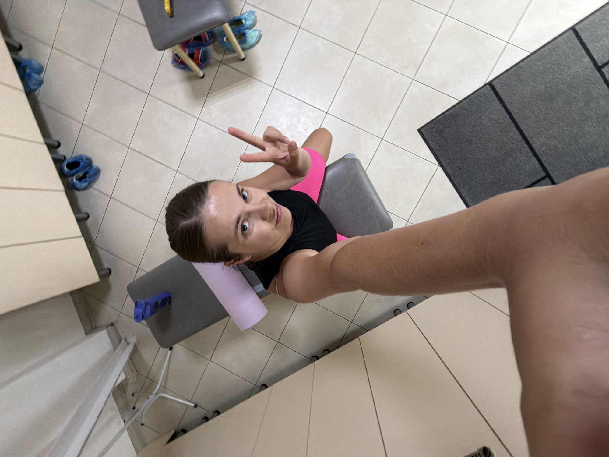 In this selfie taken from a high angle, a woman with her hair pulled back is seated on a gray padded bench, holding up two fingers in a peace sign. She is wearing a black tank top and bright pink shorts. To her left, a light purple foam roller rests on the bench. The background shows a tiled floor, with some blue shoe covers and other footwear visible in the upper left corner. A dark gray mat is present on the floor to the right. The overall setting appears to be an indoor facility, possibly a gym or a therapy room, with a clean and sterile environment. The lighting suggests it is daytime. The woman's expression is one of playful engagement with the camera, indicating a positive or lighthearted mood. There is no visible text in the image.
