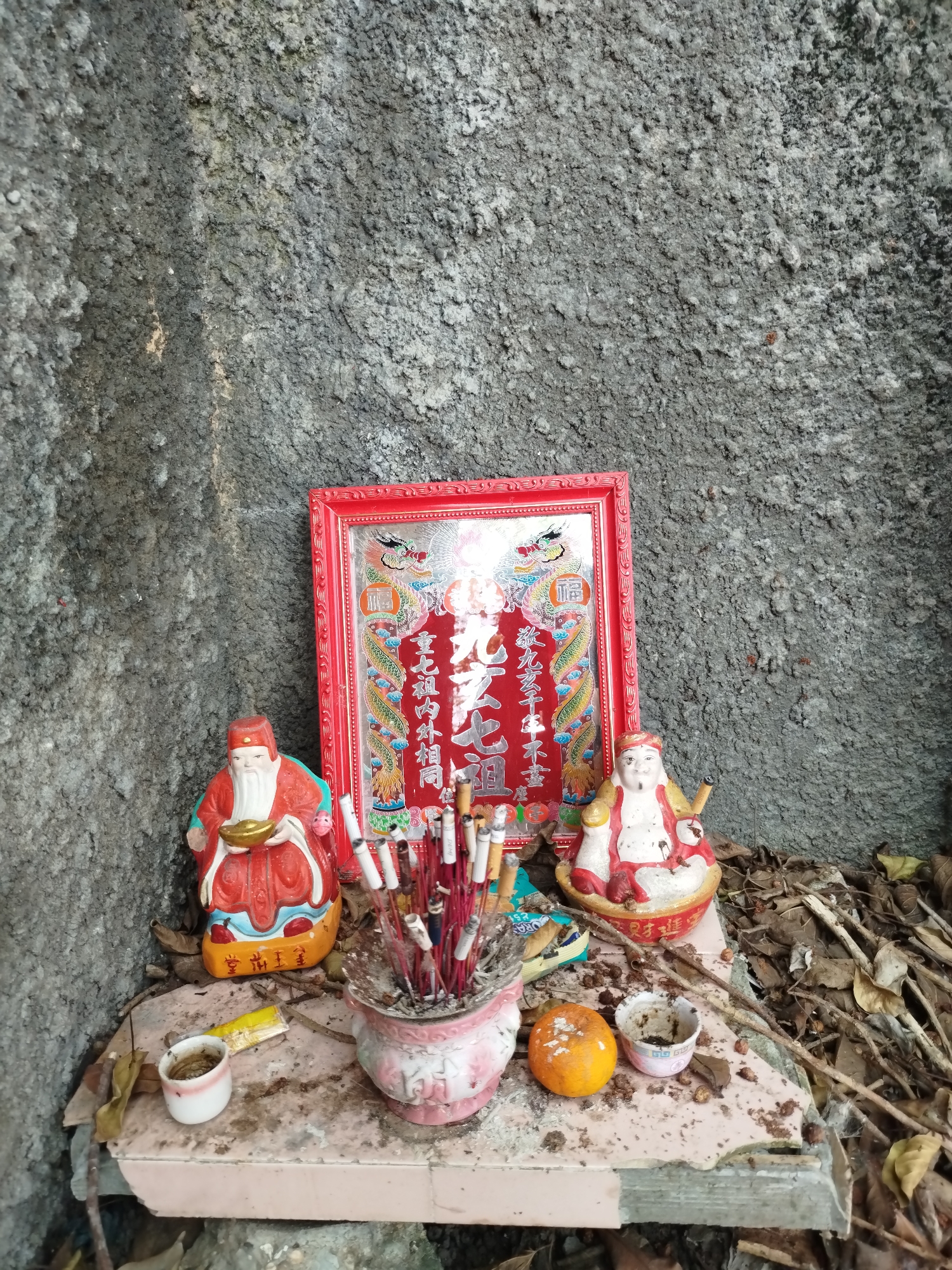 Here is a detailed description of the image:
**Location:** The photo likely depicts a small, informal shrine located outdoors. While it's difficult to pinpoint the exact location, the general architectural style and presence of a shrine suggests a location in Ho Chi Minh City, Vietnam, or a similar Southeast Asian urban area.
**Environment:** The shrine is set against a rough, gray concrete wall. Debris, leaves, and twigs are scattered around the base of the shrine, indicating an unkempt or neglected area.
**Objects:**
* **Frame:** A red frame with decorative dragon motifs holds a print with Chinese characters.
* **Statues:** Two small, painted statues, likely depicting deities or spiritual figures, flank the frame. One holds a golden bowl, and the other has a pipe.
* **Incense Burner:** A pink ceramic pot holds a bundle of incense sticks, many of which have been burnt.
* **Offerings:** A single orange and two small cups are placed on the shrine as offerings. A piece of yellow paper is also visible, possibly a talisman or prayer note.
* **Platform:** The objects are arranged on a small, possibly broken, pink-tiled platform.
**Symbolism/Interpretation:**
The scene suggests a small, personal shrine where someone has been offering prayers or making requests. The deities, frame, and offerings point to a blend of traditional Chinese and local Vietnamese religious practices. The presence of burnt incense indicates recent use of the shrine. The unkempt environment might suggest that the shrine is either abandoned or located in a low-traffic area.